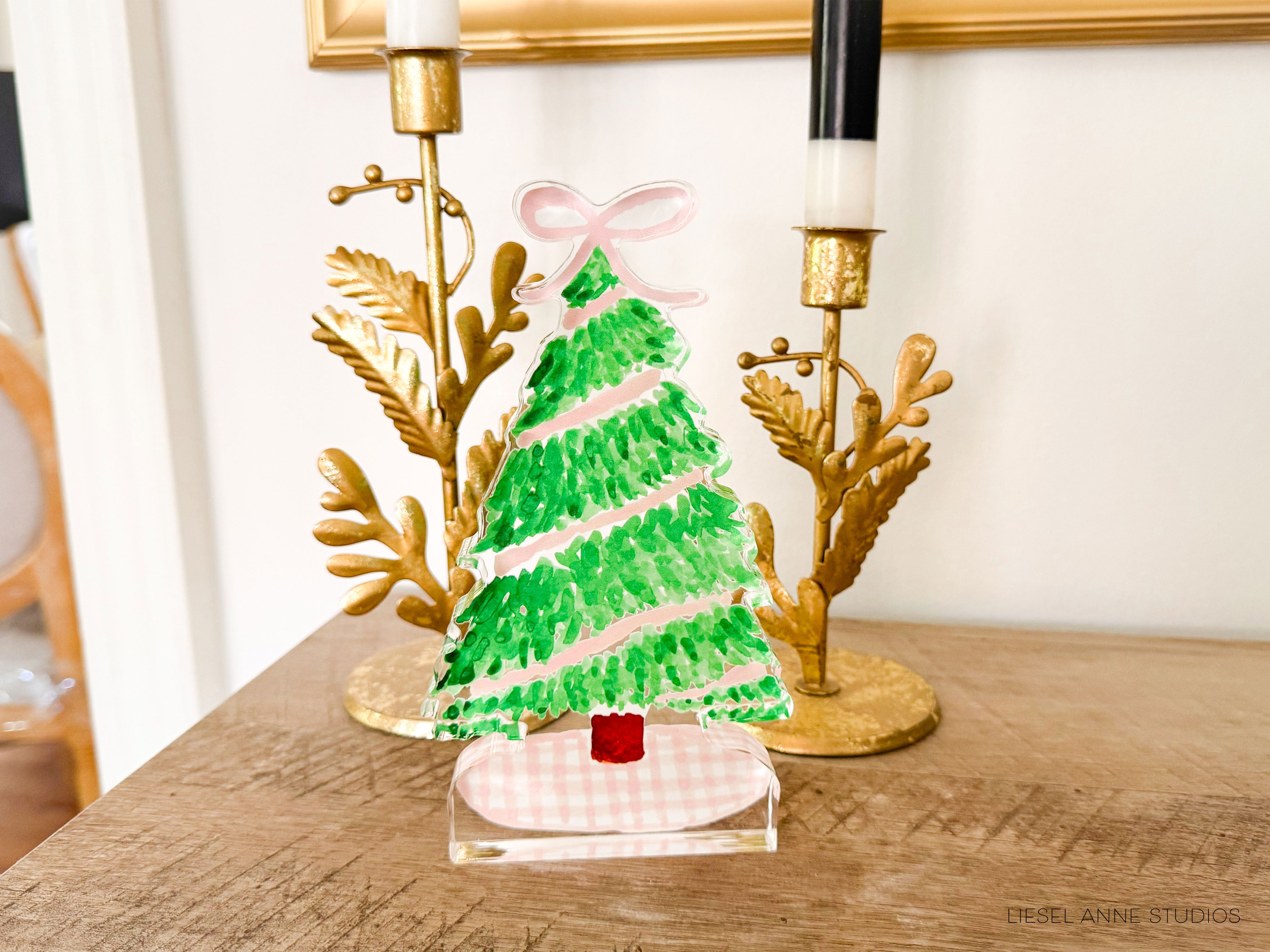 A green and pink painted Christmas tree on a wooden table, with two gold candlesticks and a gold-framed picture in the background.