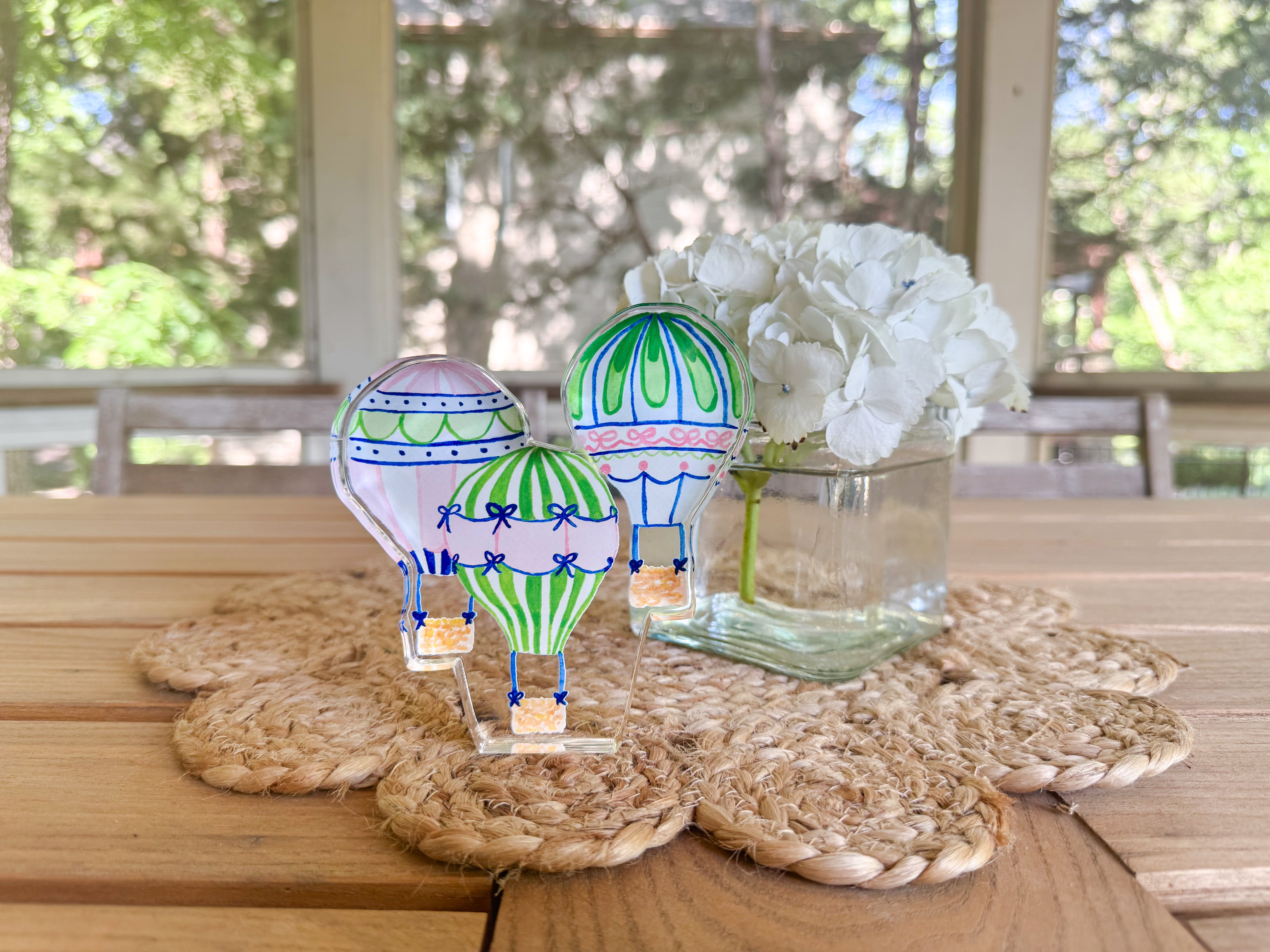 a wooden table with three colorful hot air balloons, a vase of white flowers, and a woven placemat.