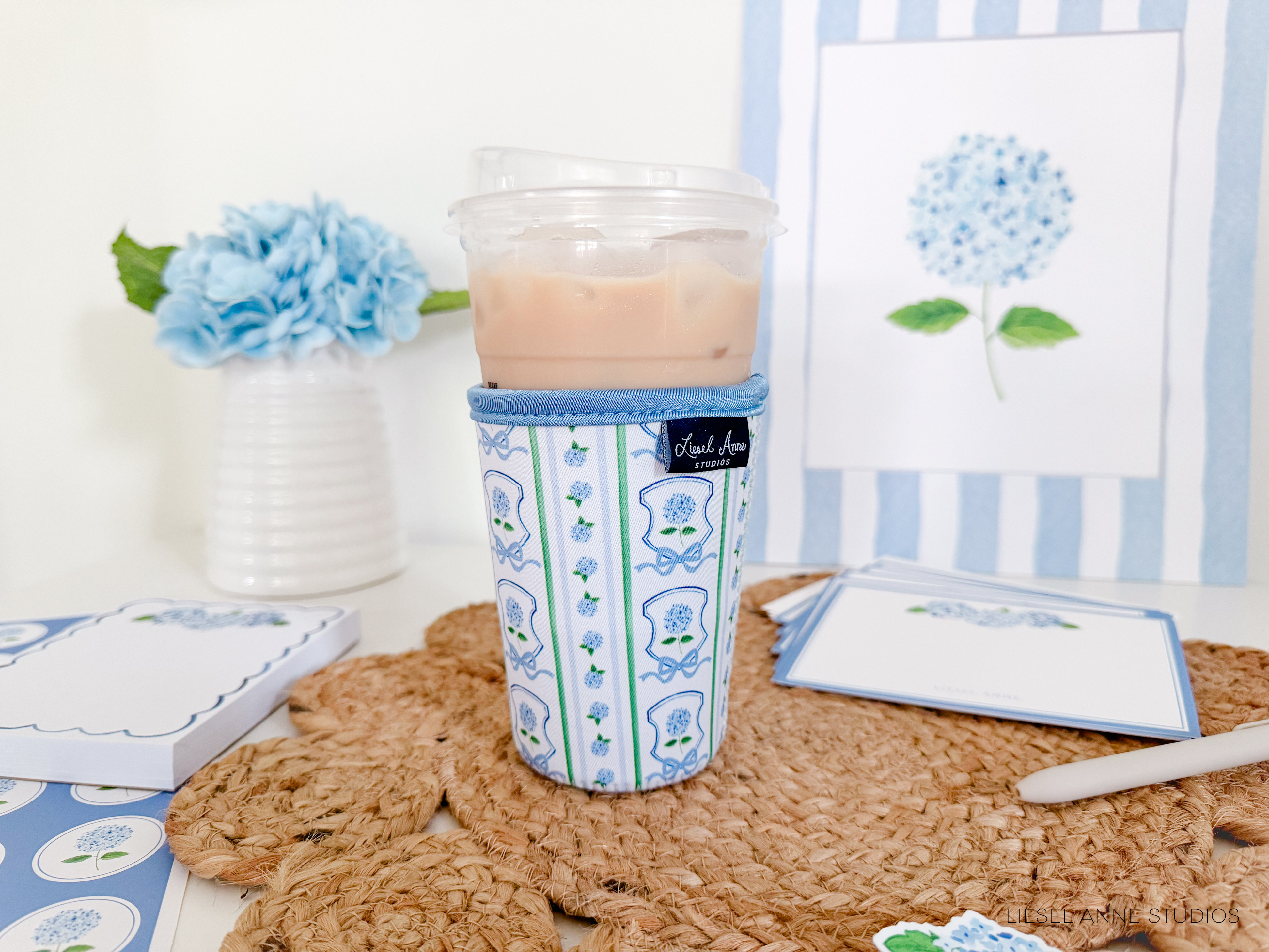 a cup of iced tea with a blue and white floral pattern, placed on a woven placemat. The cup has a lid and is accompanied by a pen and a stack of blue and white cards. In the background, there is a vase with blue flowers, and a blue and white striped picture frame.