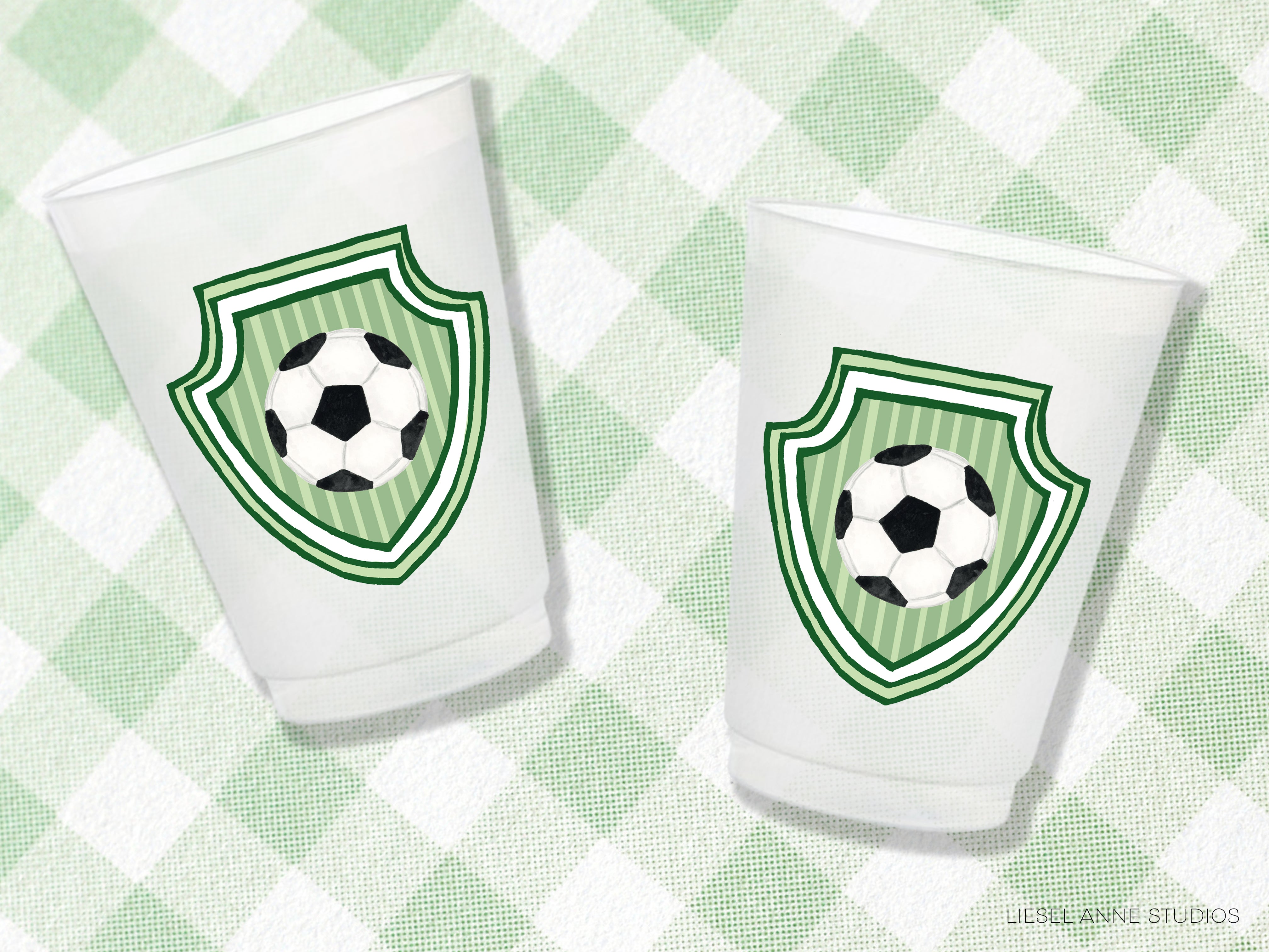 Two white plastic cups with green and white soccer ball designs on them, placed on a green and white checkered background.
