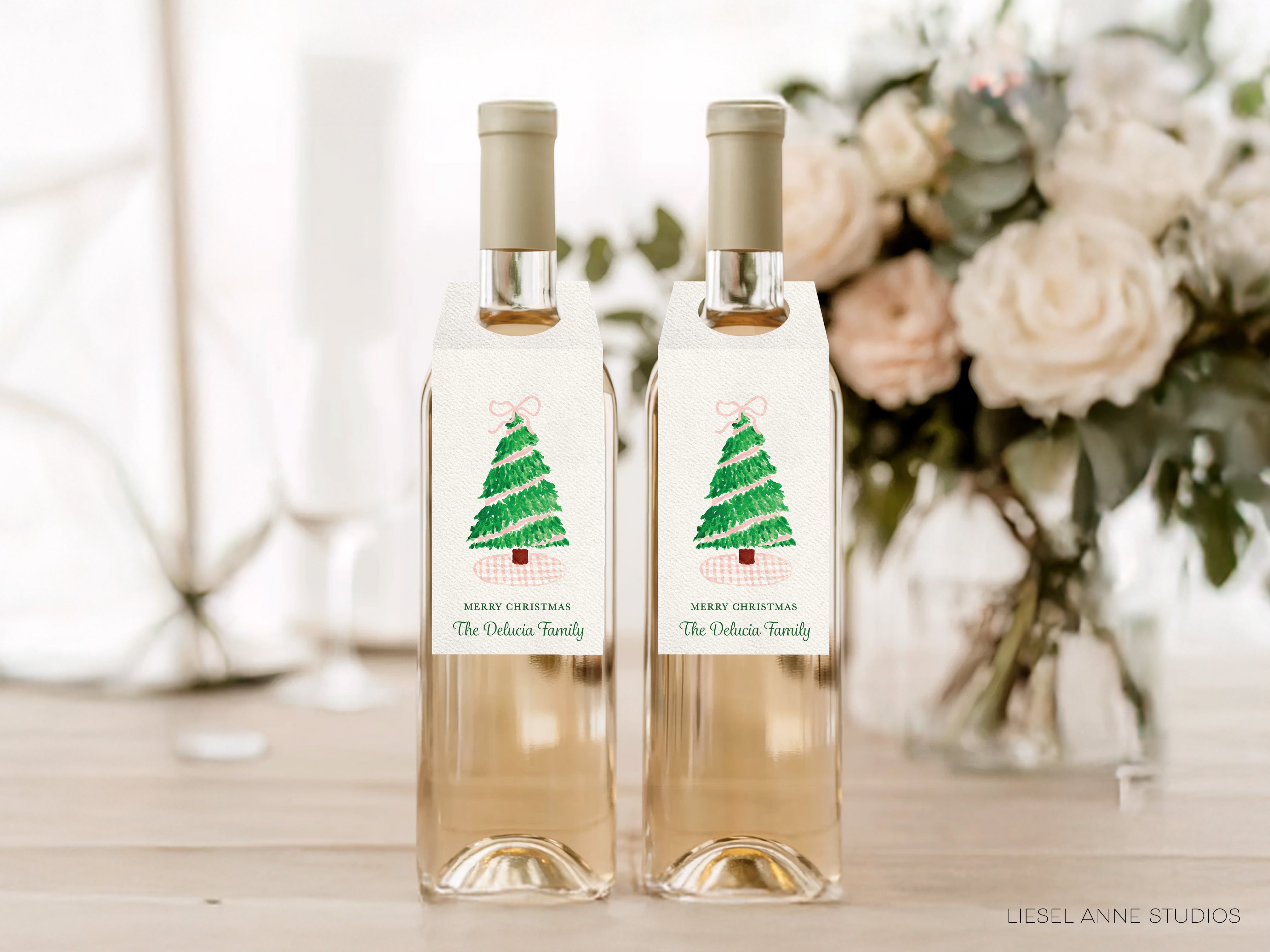 Two bottles of wine with labels that read "Wish you a Merry Christmas The Delicacies Family" are displayed on a wooden table, with a vase of white flowers in the background.