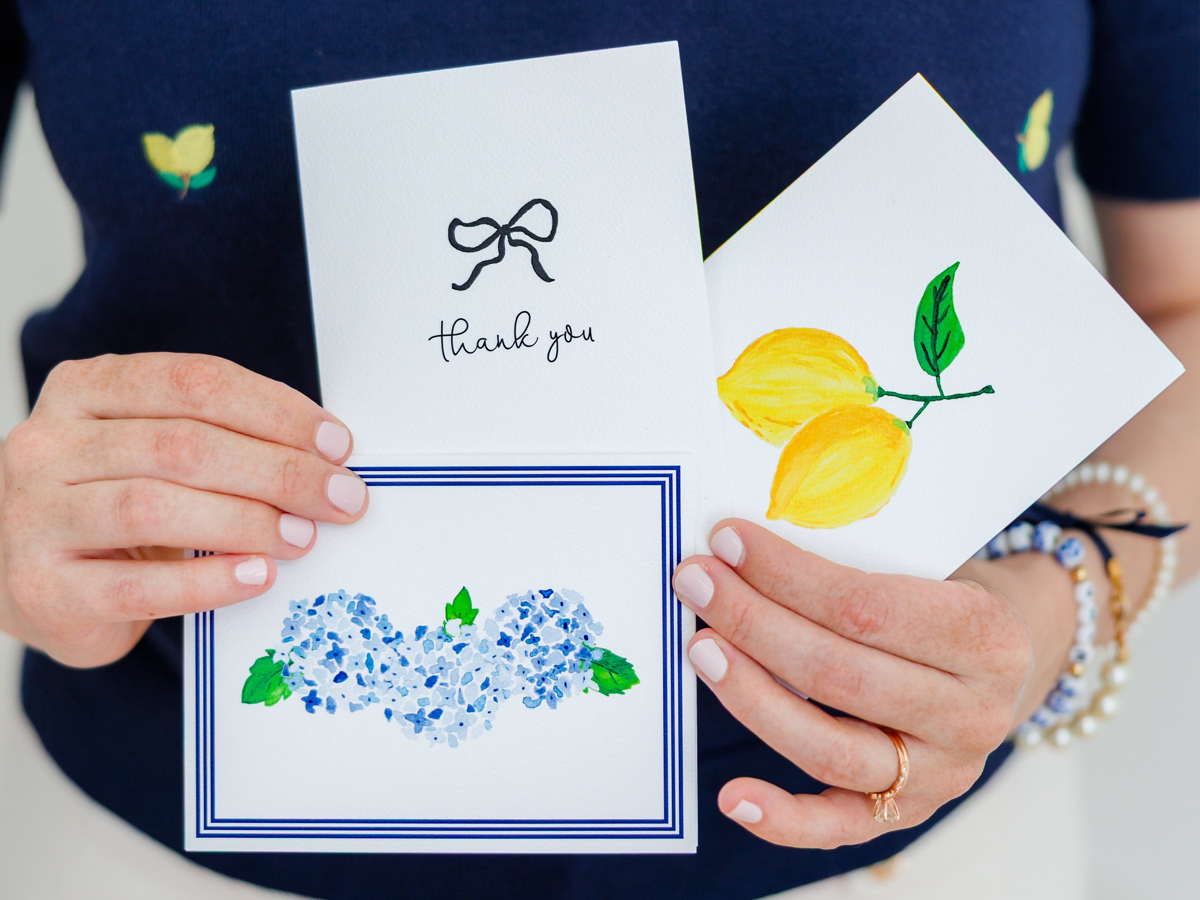 A person is holding three greeting cards, each with different designs and text. The cards include a floral design, a lemon design, and a "Thank you" card.