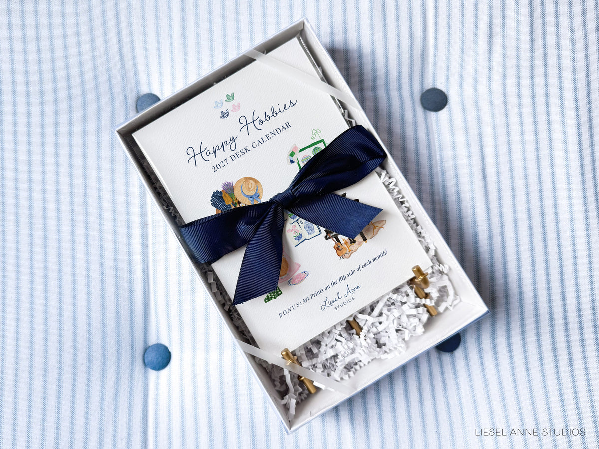 Happy Hobbies 2027 watercolor desk calendar gift box with a navy ribbon and gold easel, ready to give