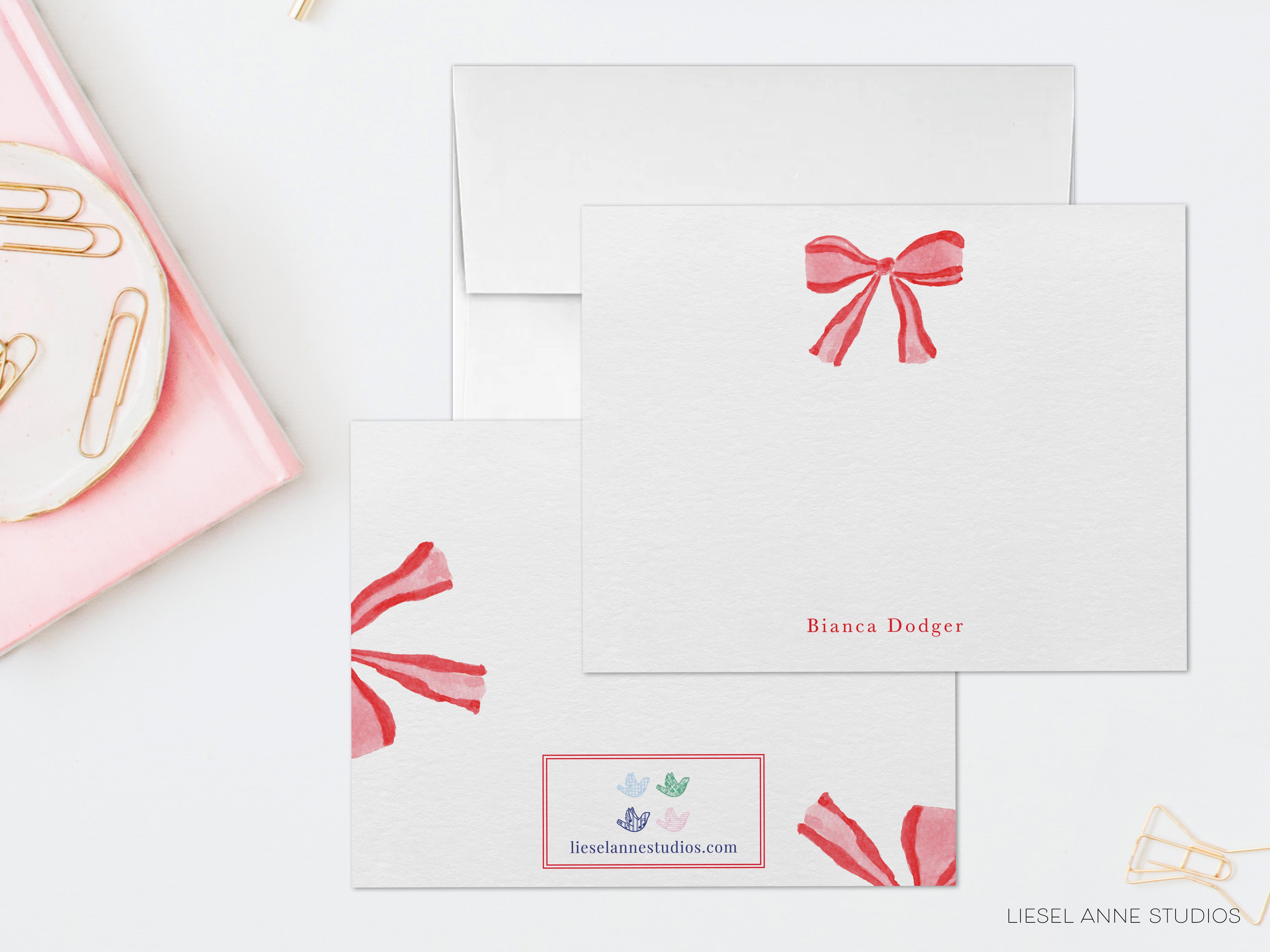a set of three white envelopes with red bows, each with a personalized name printed on them. The envelopes are arranged on a white surface, with a pink plate and some gold clips nearby.