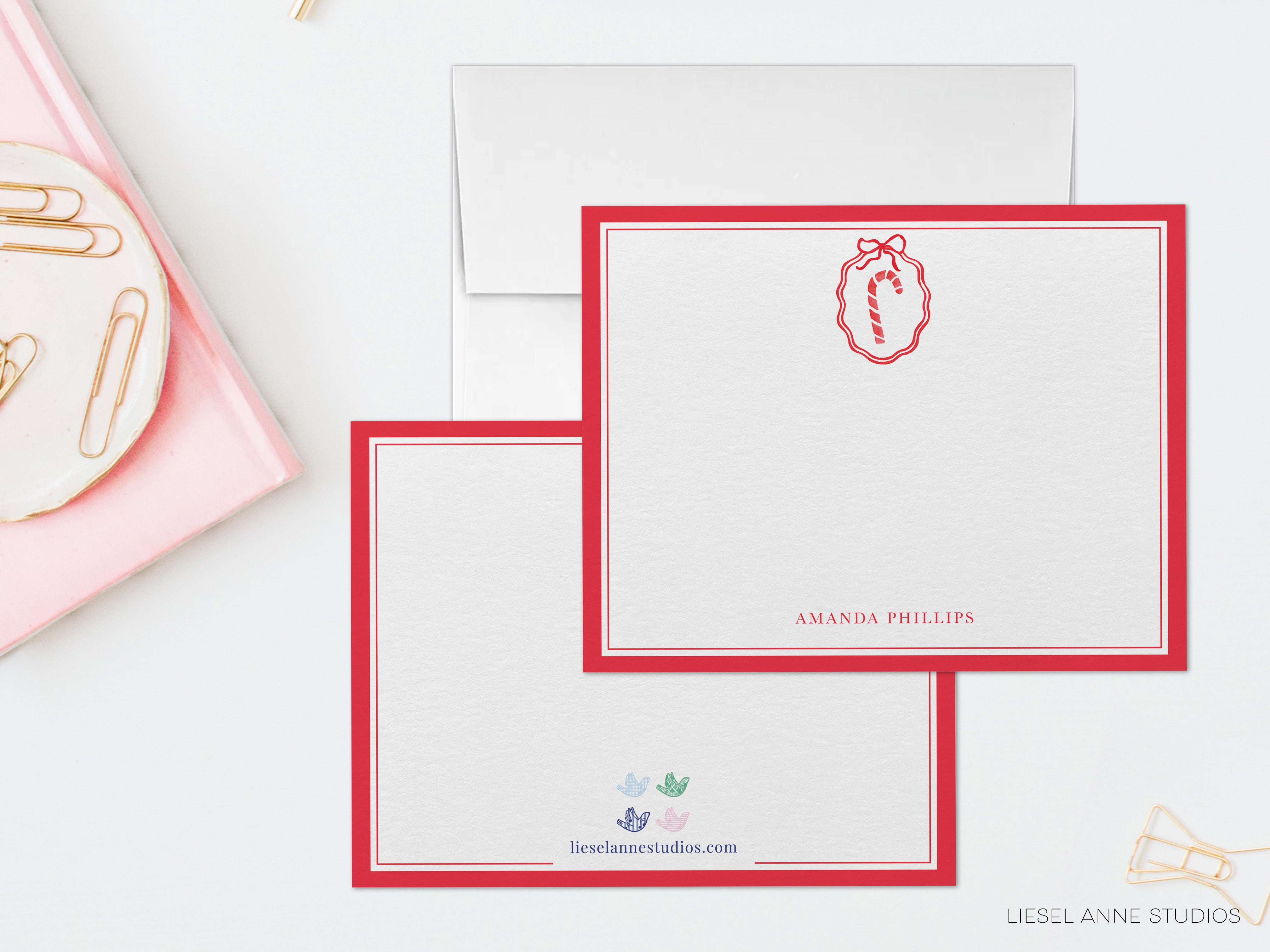 two blank red and white stationery cards with a personalized name and a red ribbon design, placed on a white surface alongside a pink plate with gold clips.