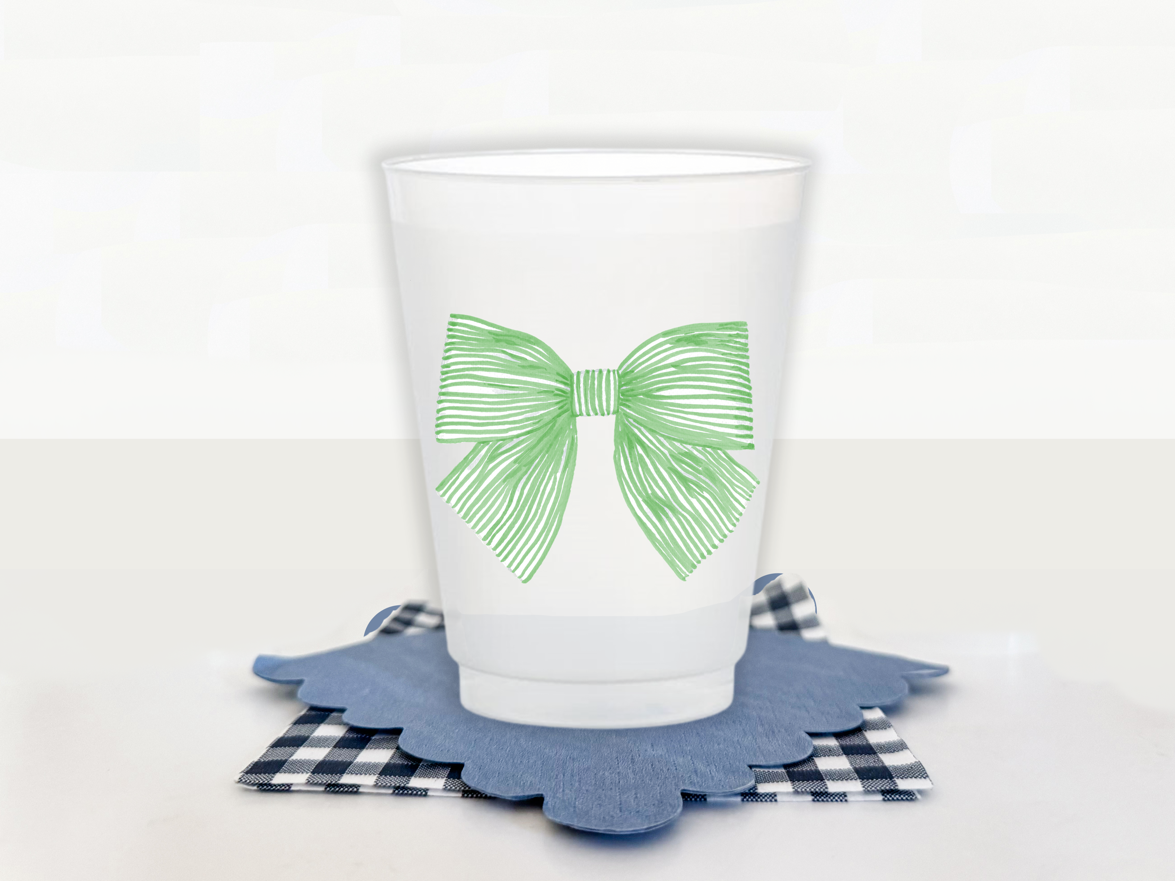 A white plastic cup with a green bow design on it, placed on a blue and white checkered coaster.