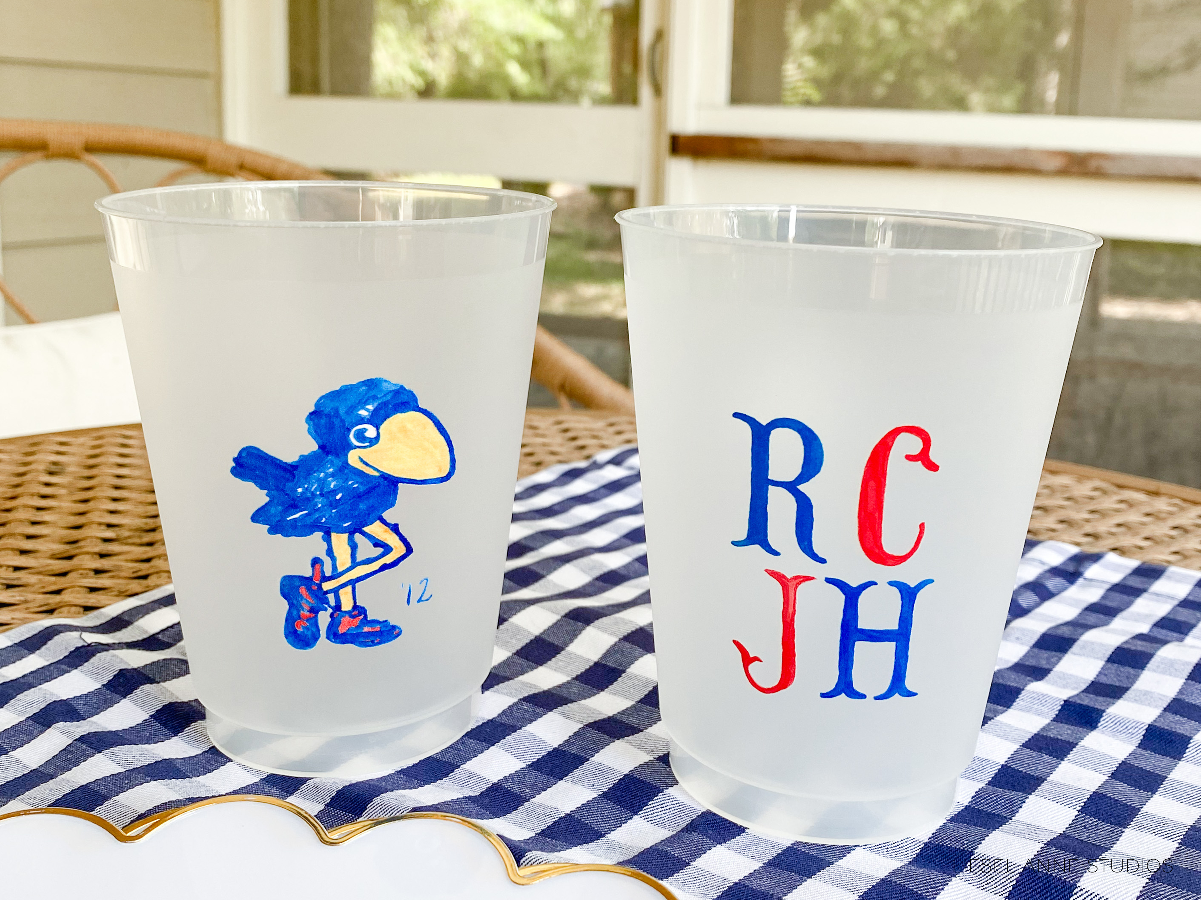 Two plastic cups with personalized monogrammed designs on them, placed on a checkered tablecloth.