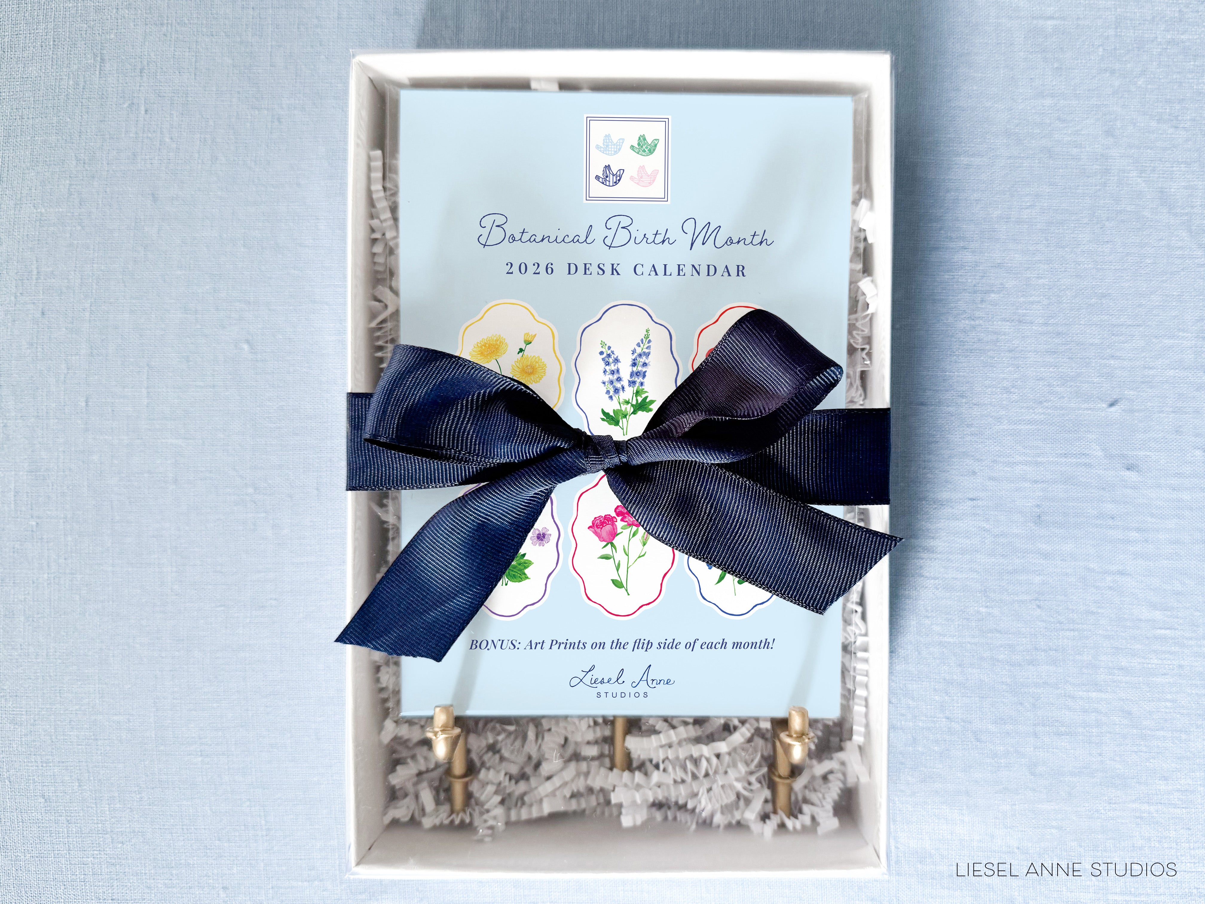 a 2020 desk calendar with a blue background and a white ribbon tied around it. The calendar is presented in a white box with a blue ribbon, and there is a small note that reads "BONUS: Art prints on the tip of each month!" The text "Liesel Anne Studio" is also visible, indicating the source or creator of the calendar.