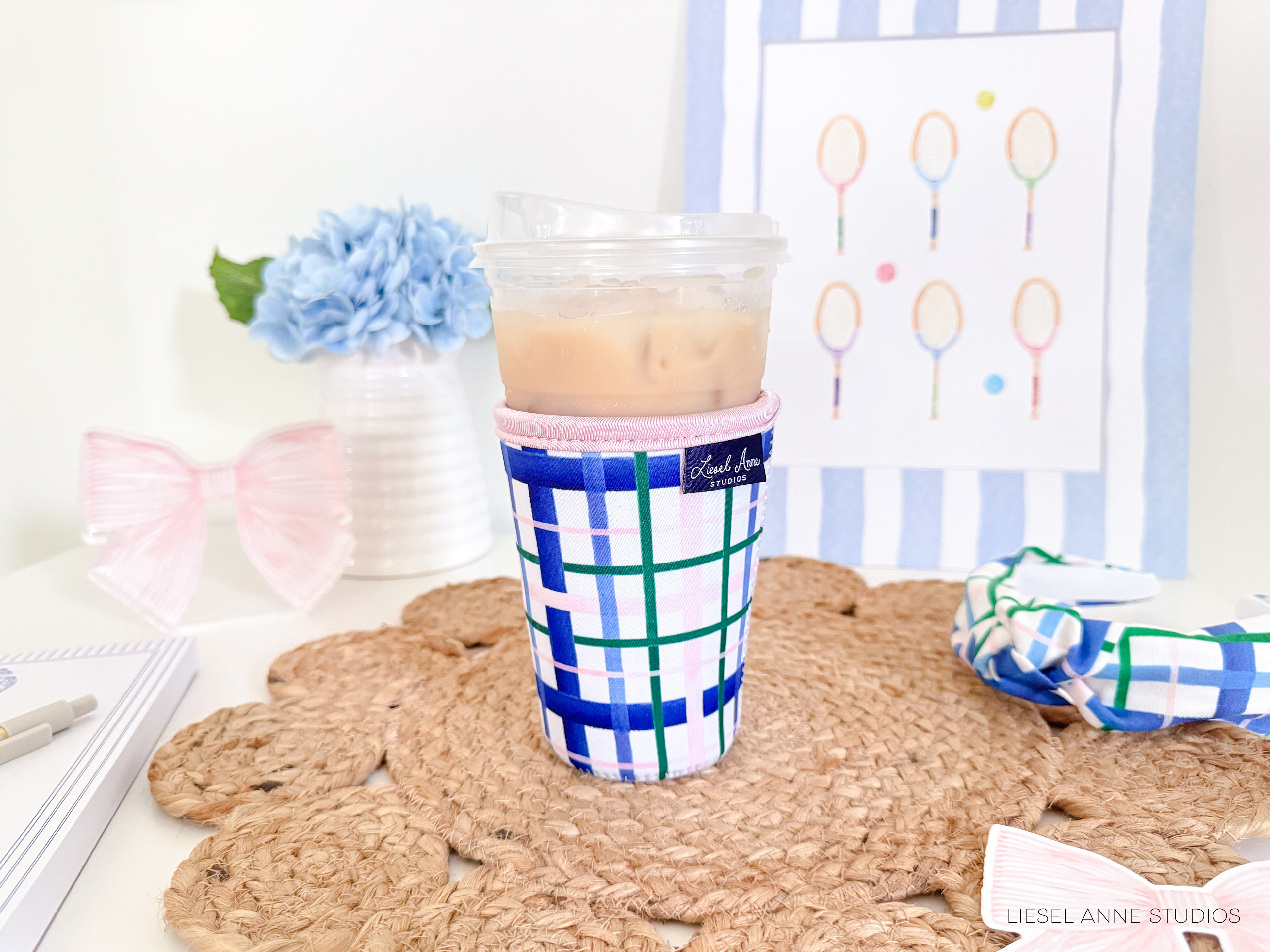 a cup of iced tea with a colorful cup warmer, placed on a woven placemat. In the background, there is a framed picture with tennis rackets, a vase with a blue flower, and a pink bow.