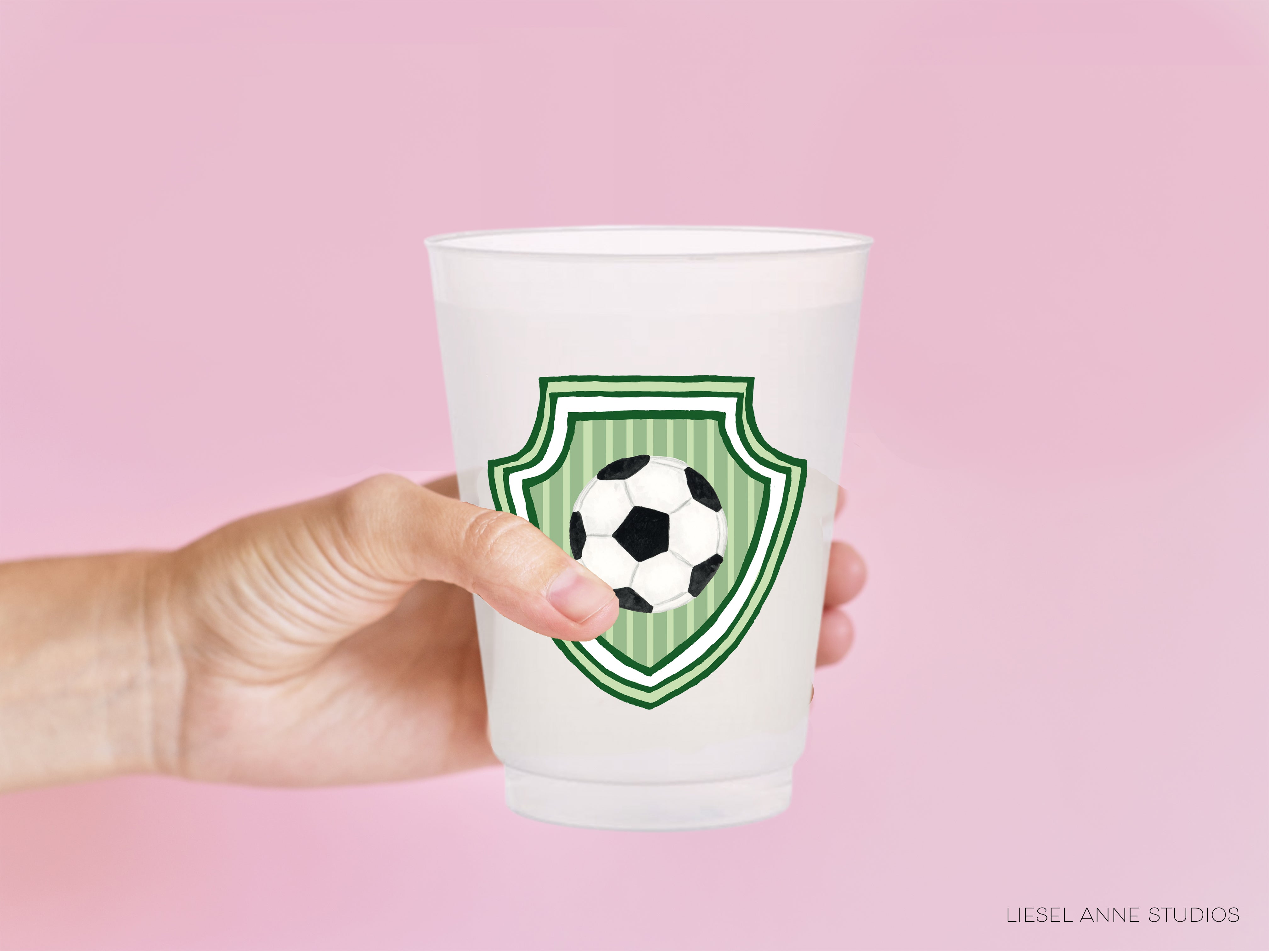 A hand holding a white plastic cup with a soccer ball design on it against a pink background.