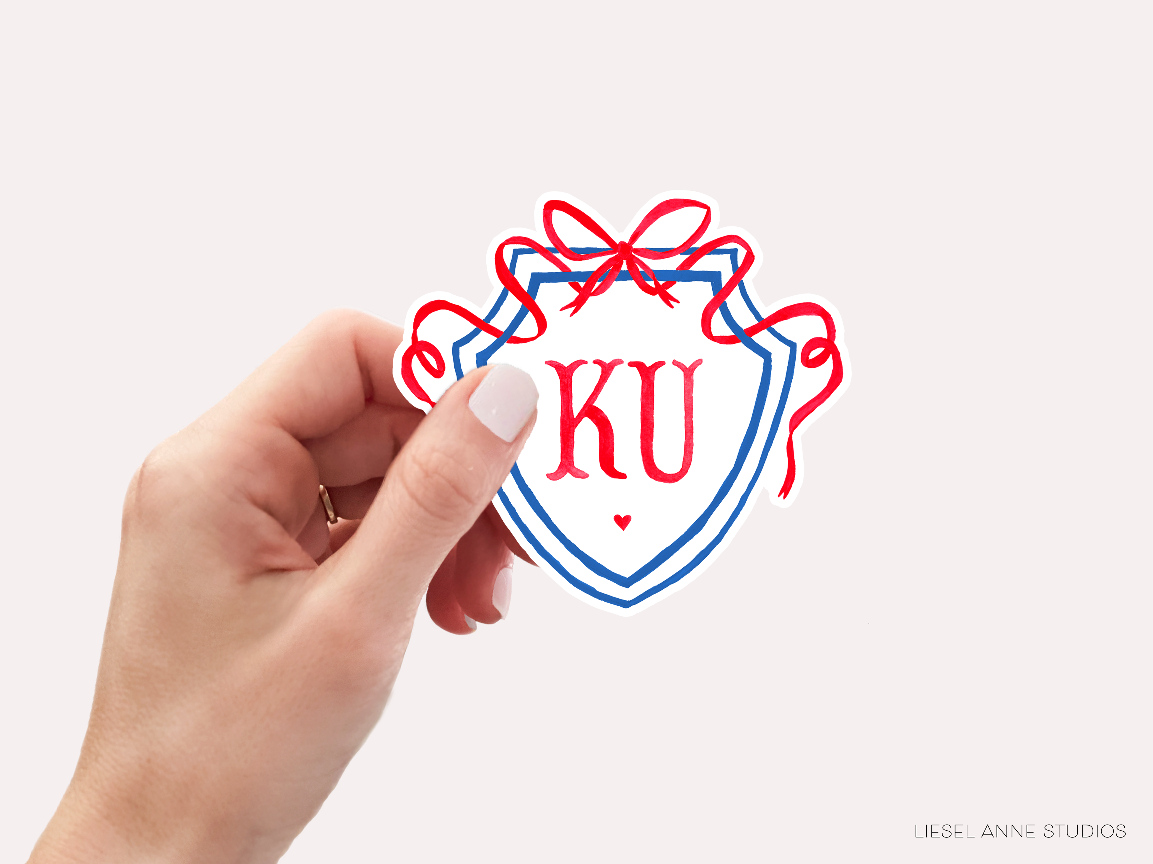 A hand holding a shield-shaped sticker with the initials 'KU' and a red ribbon design.