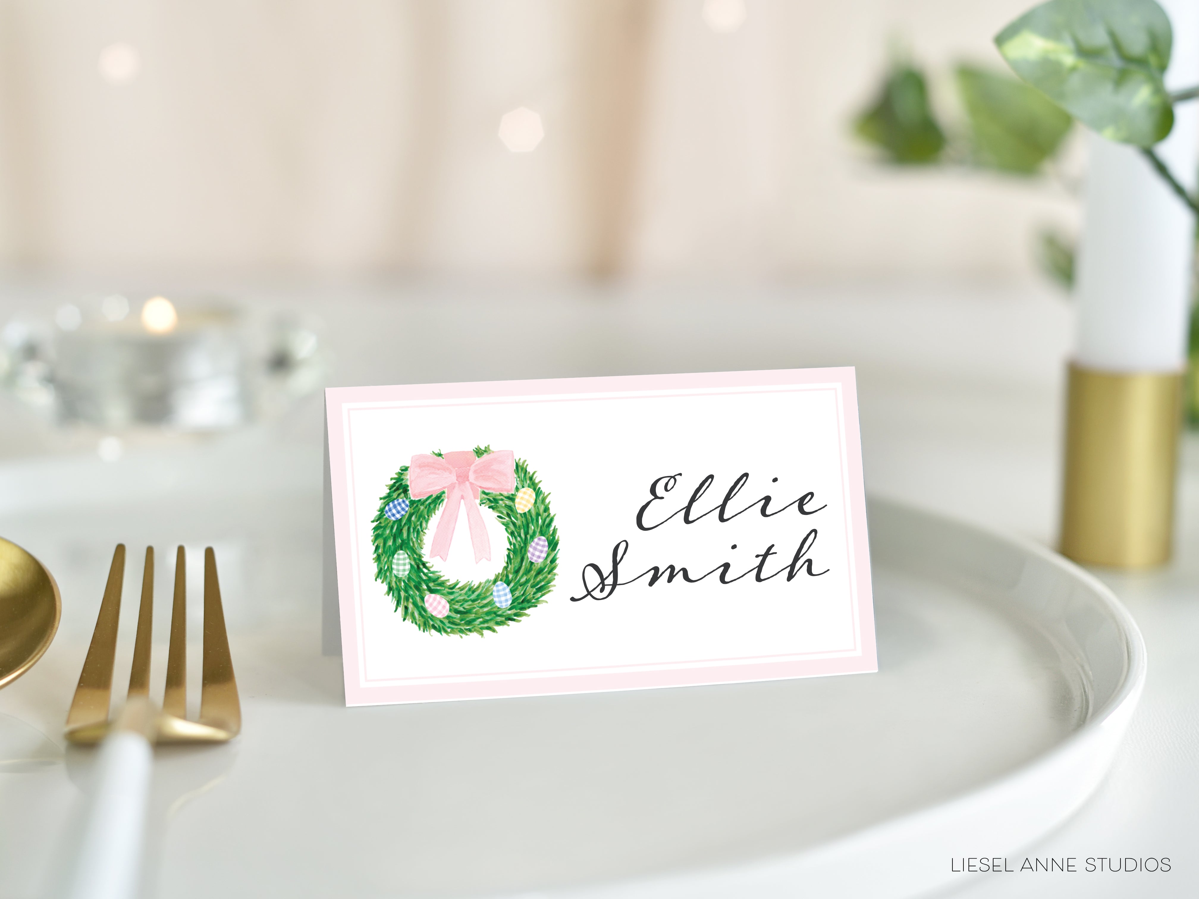 A pink name card with a wreath design and the name "Ellie Smith" written on it, placed on a white plate.