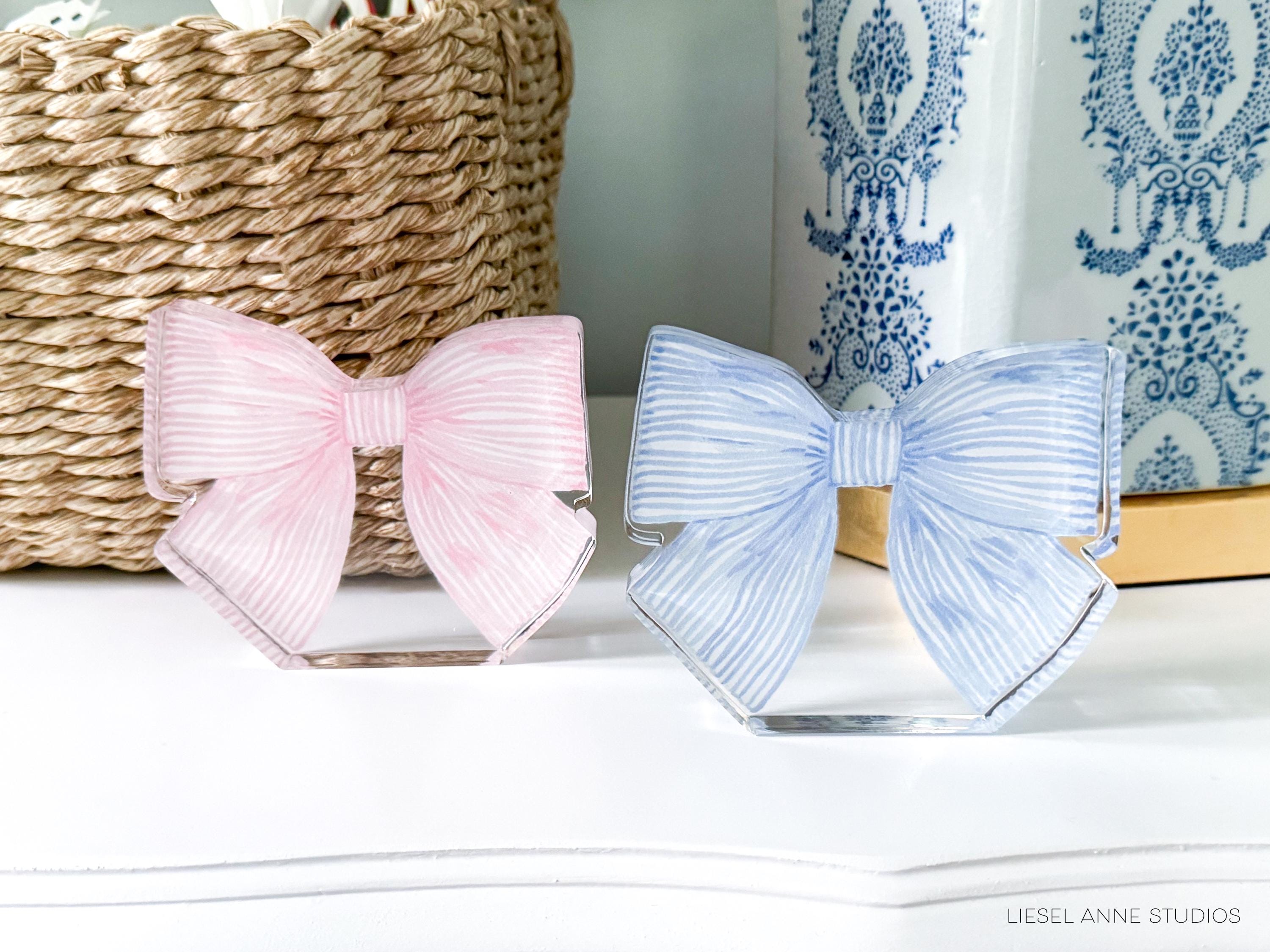 two decorative bow ties, one pink and one blue, placed on a white surface. There is also a woven basket and a blue and white patterned vase in the background.