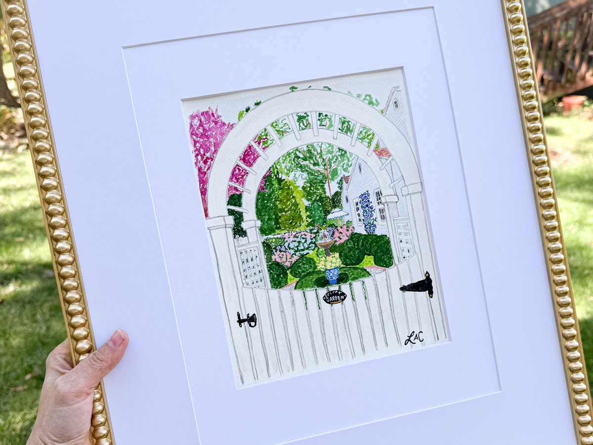 A hand holding a framed painting of a white gate with a colorful garden scene behind it.
