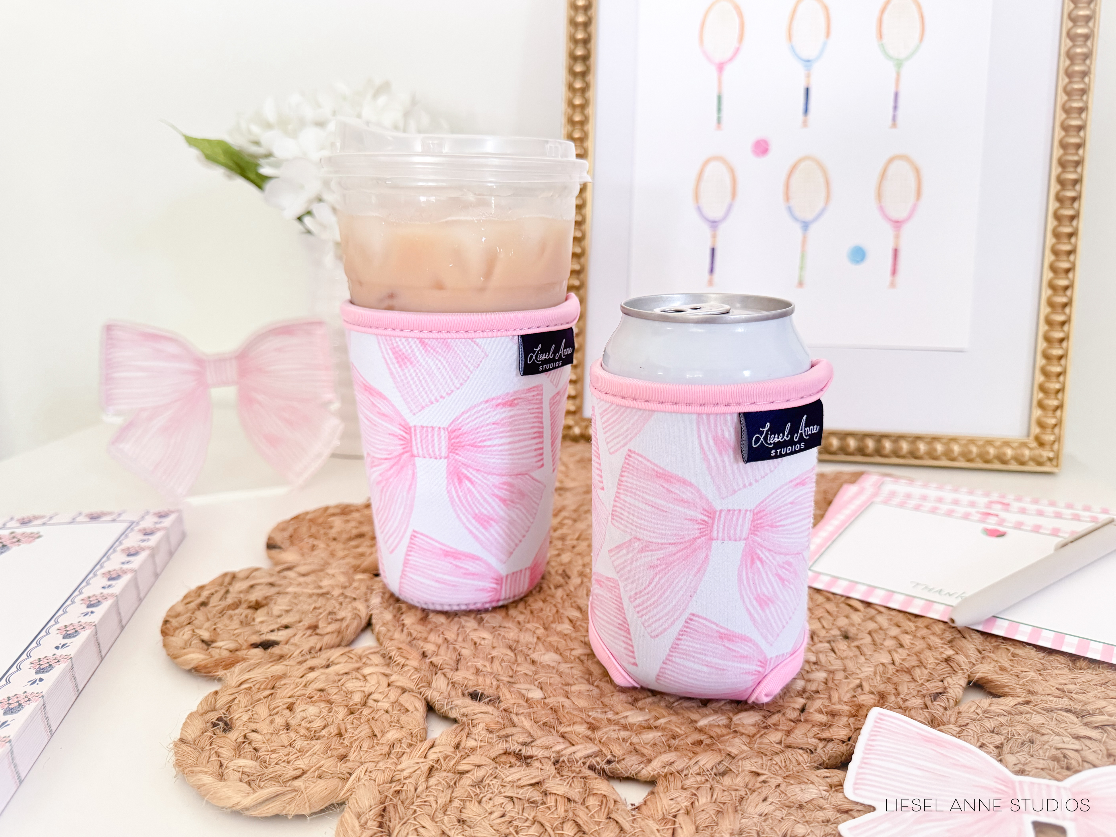 two pink bow-themed drink holders, one larger and one smaller, placed on a woven placemat. In the background, there is a framed picture with tennis racket illustrations.