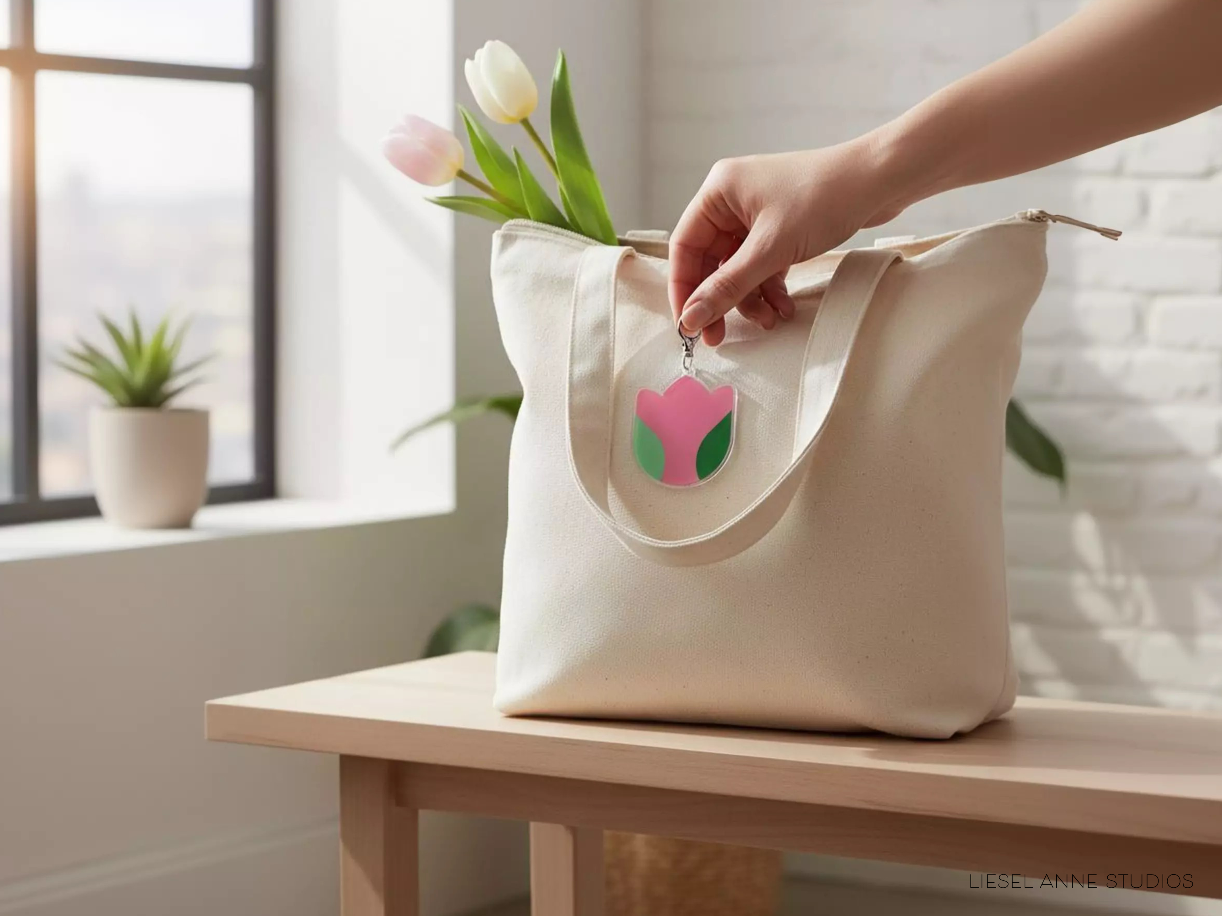 A hand is holding a white tote bag with a pink and green flower charm attached to it, placed on a wooden table near a window.