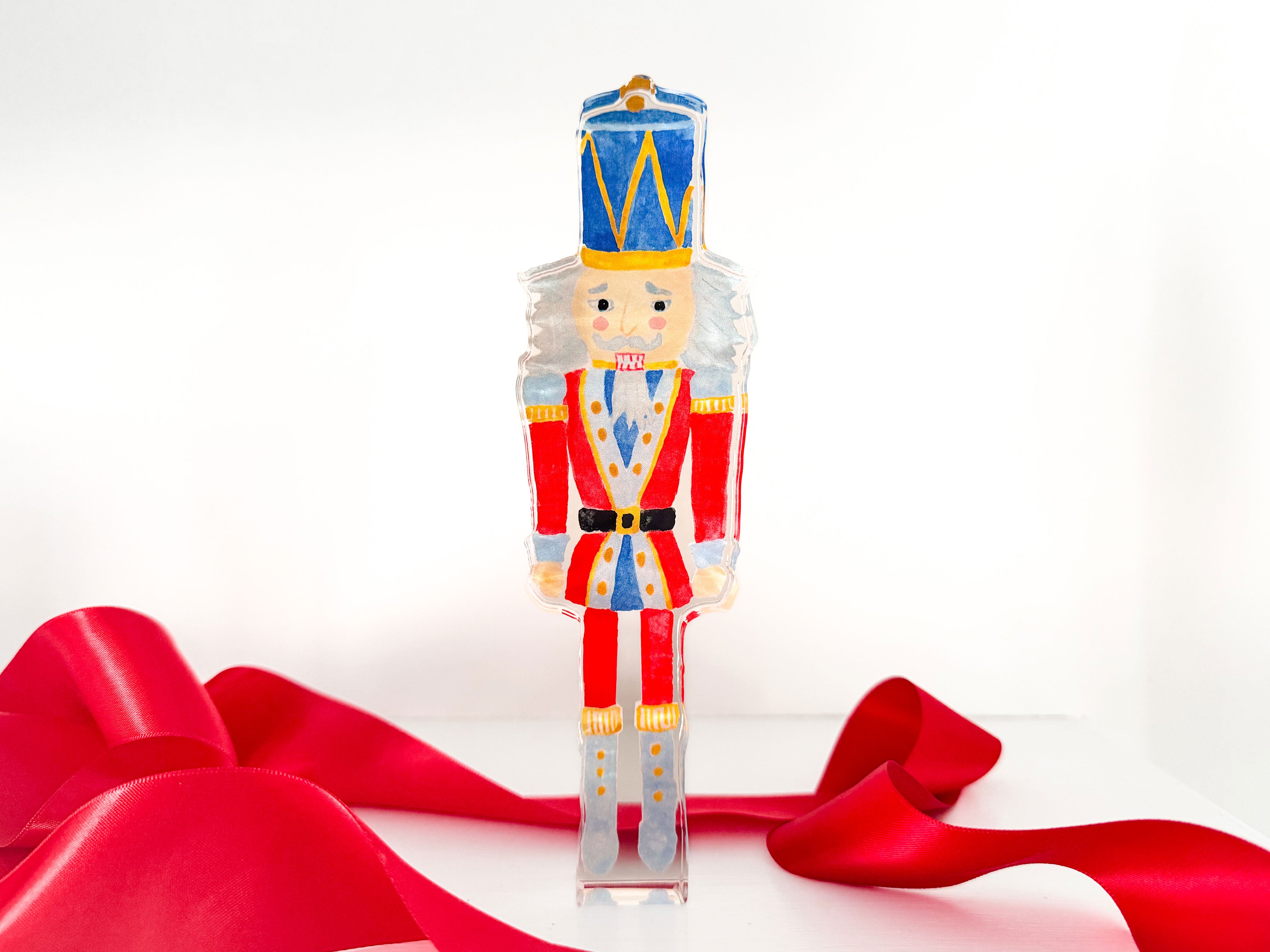 A clear plastic nutcracker figure with a blue hat and red and gold outfit stands on a red ribbon against a white background.