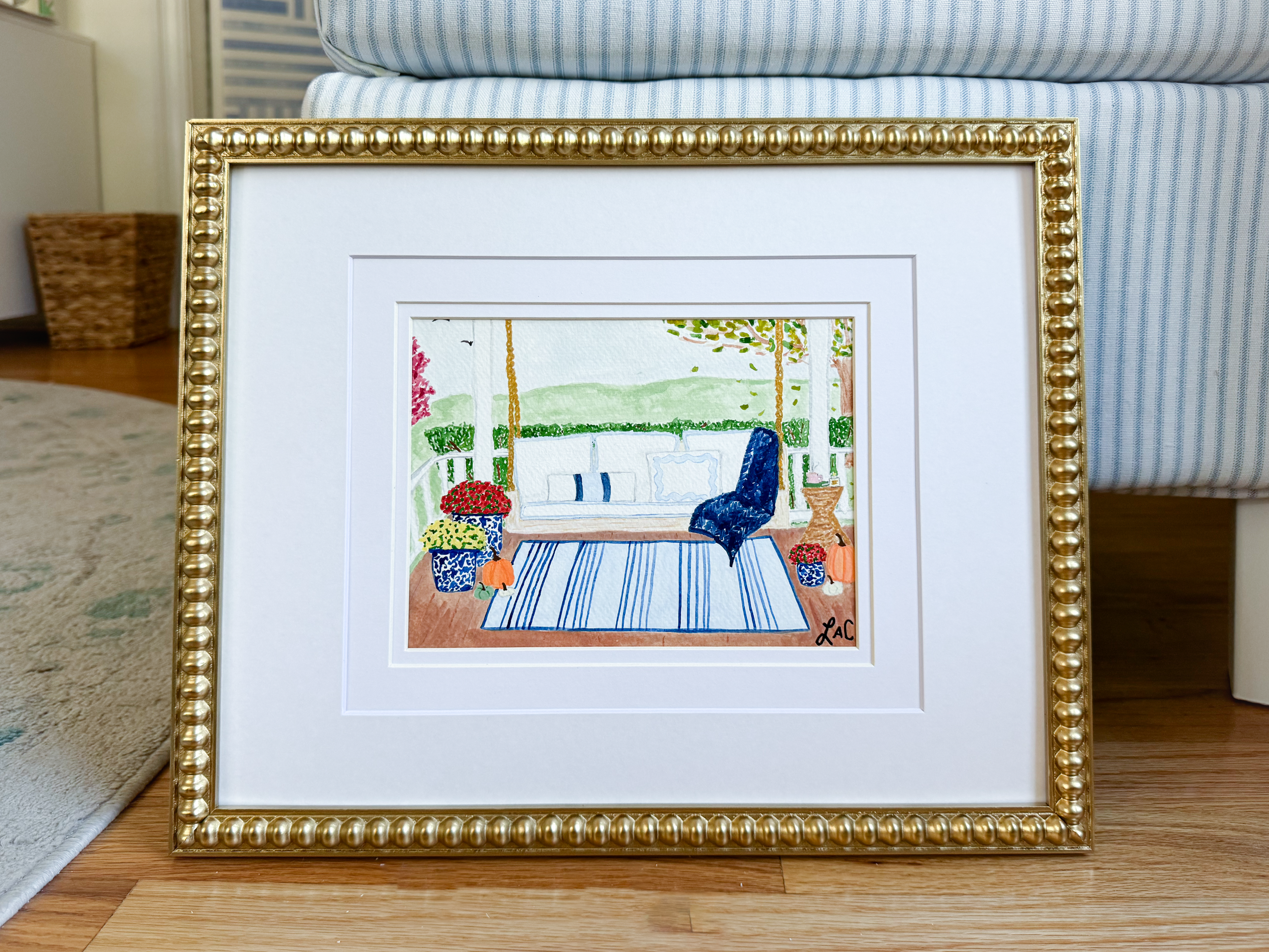 a framed painting depicting a serene outdoor scene with a blue chair, a white railing, and a blue and white striped rug.
