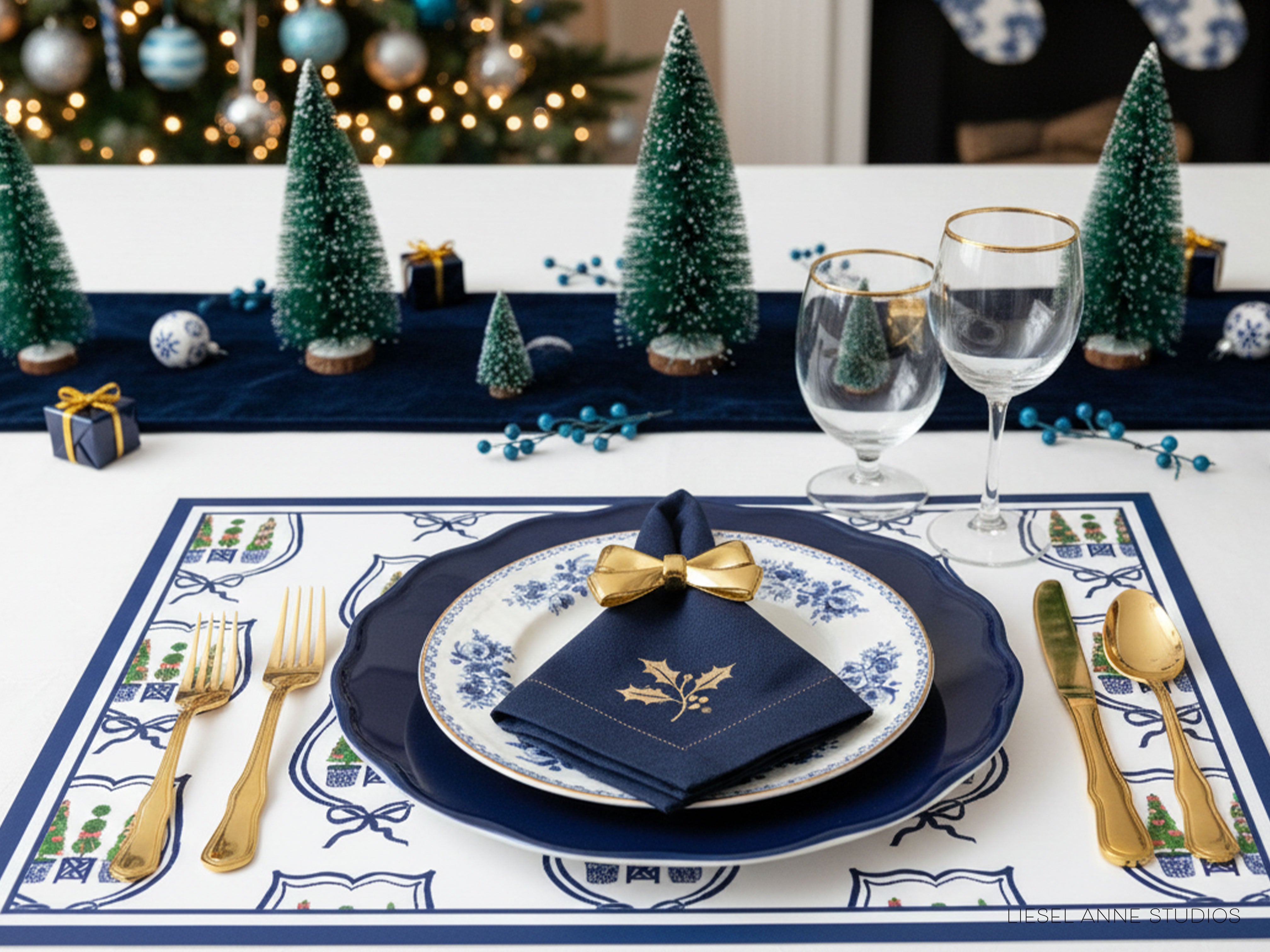 a beautifully set table for a holiday meal, with a blue and white tablecloth, gold-rimmed glasses, a blue and white plate with a gold bow, and a small Christmas tree.