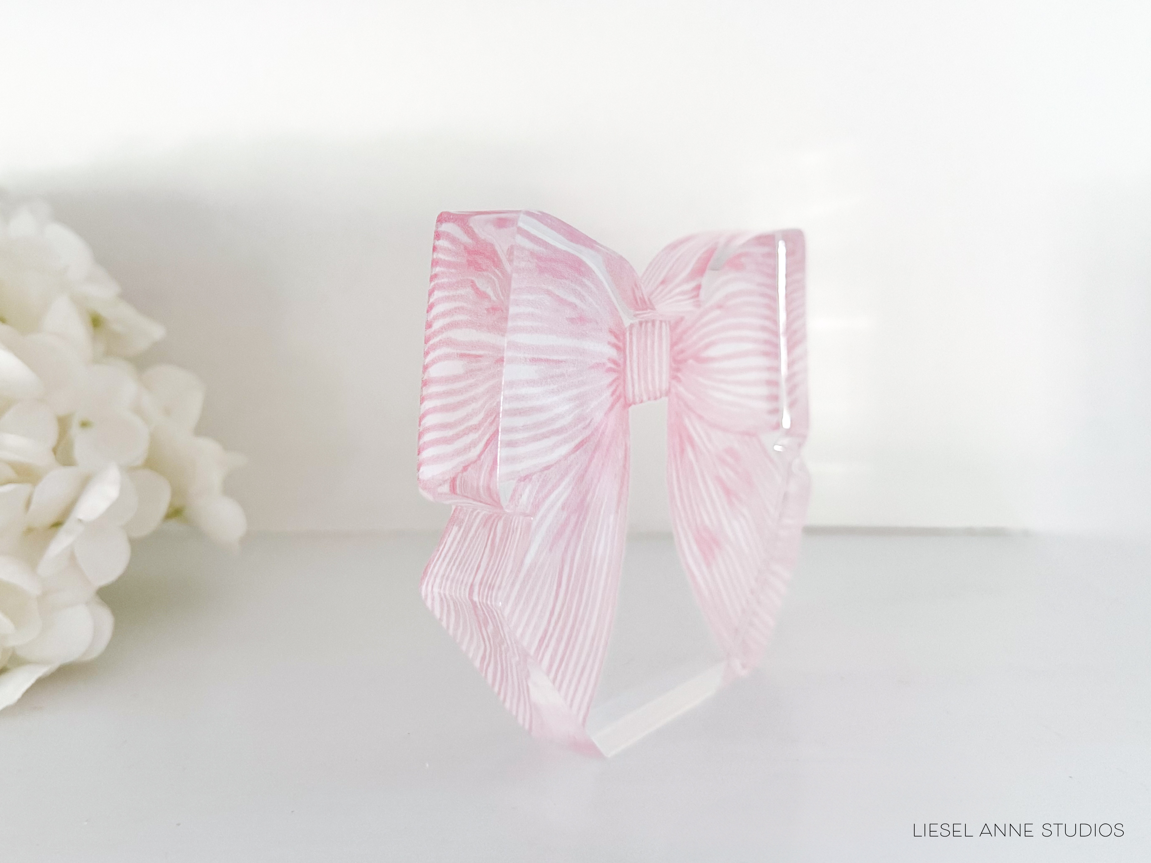 A pink bow-shaped object is displayed on a white surface, with a white flower arrangement visible in the background.