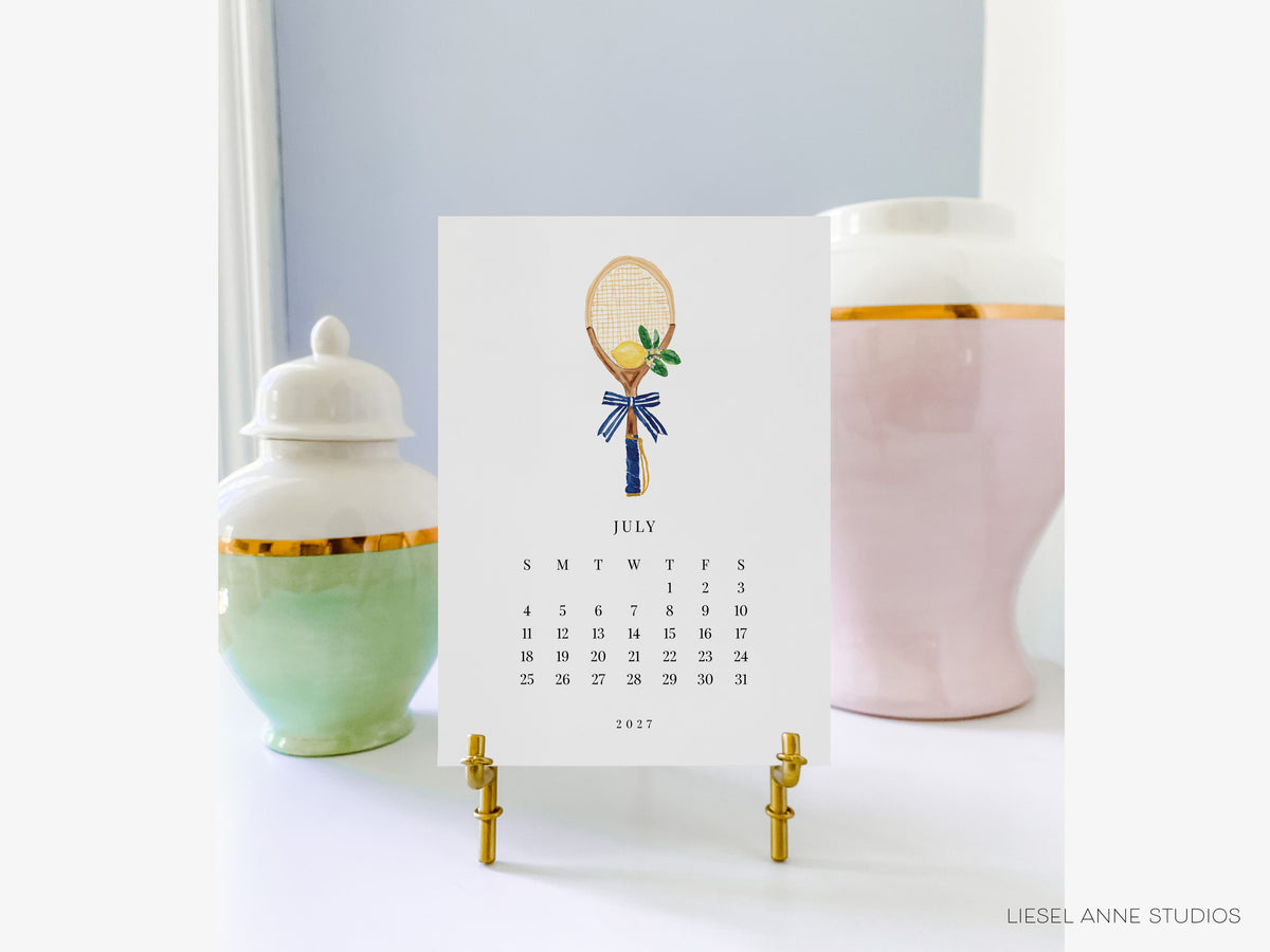 A calendar for July 2017 featuring a tennis racket illustration, placed on a stand in front of two decorative vases.
