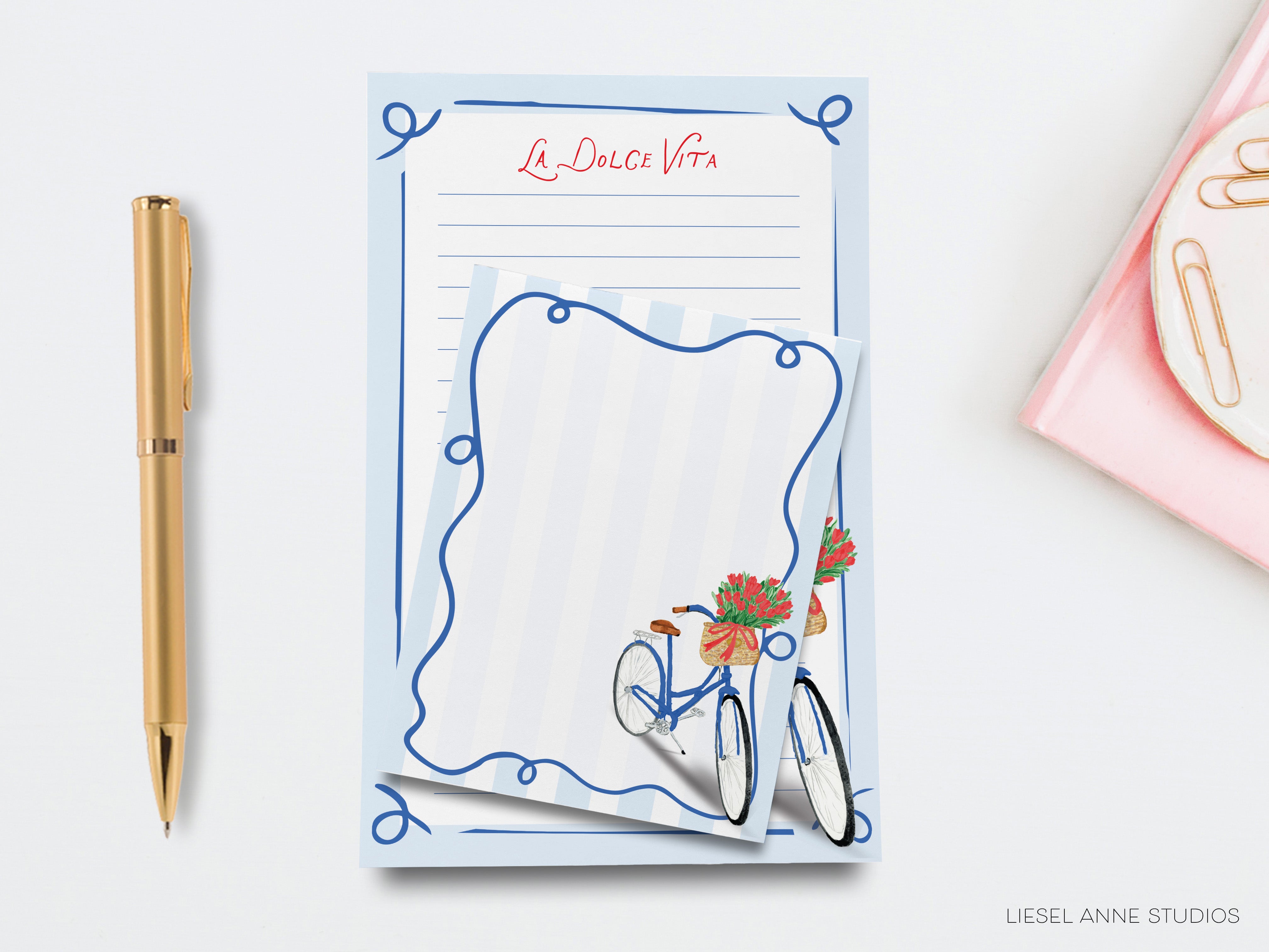 a blue notepad with a bicycle illustration, a gold pen, and a pink object on a white background.