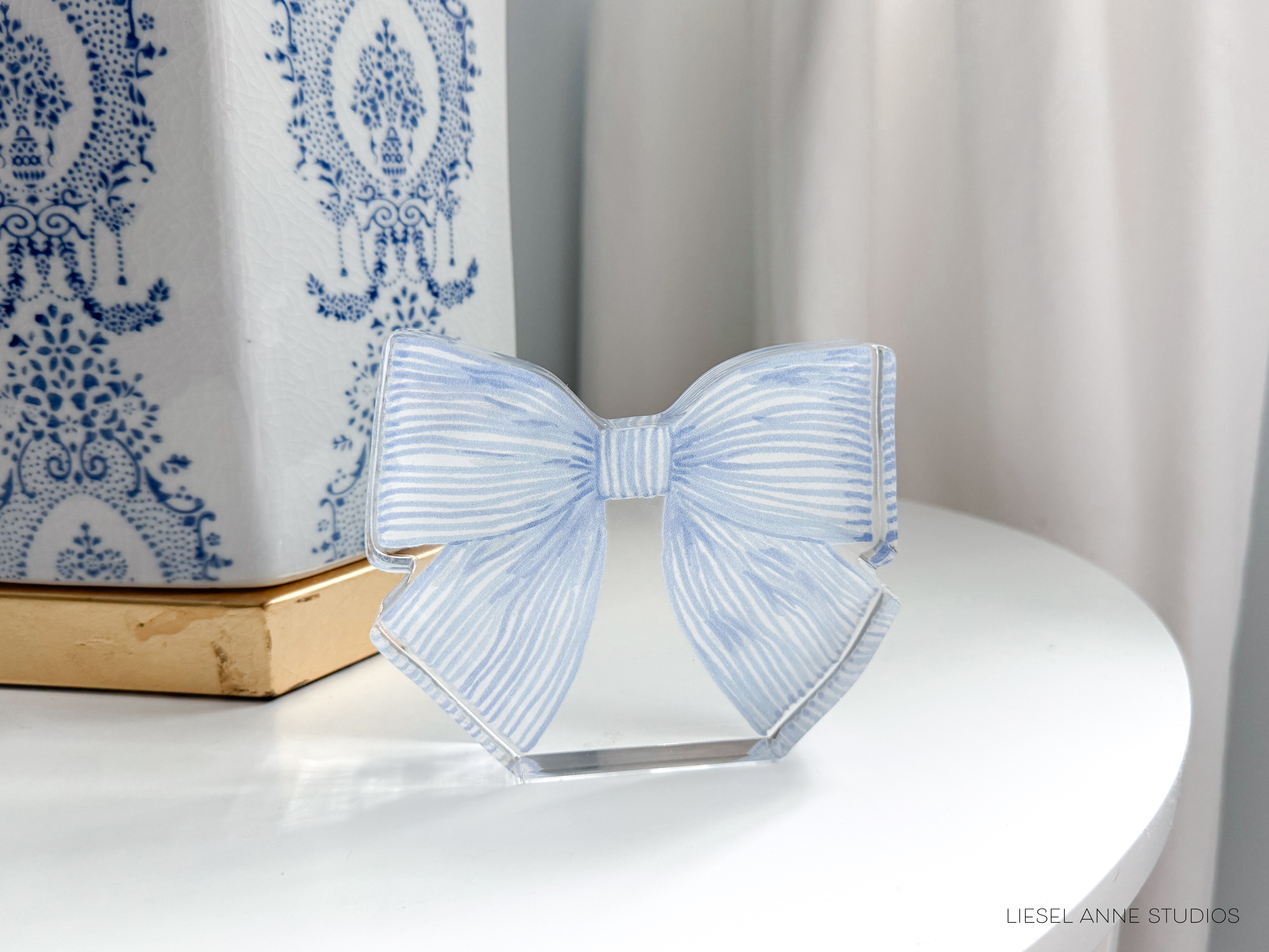 A decorative blue and white striped bow is placed on a white surface, with a white lampshade featuring a blue and white floral pattern in the background.