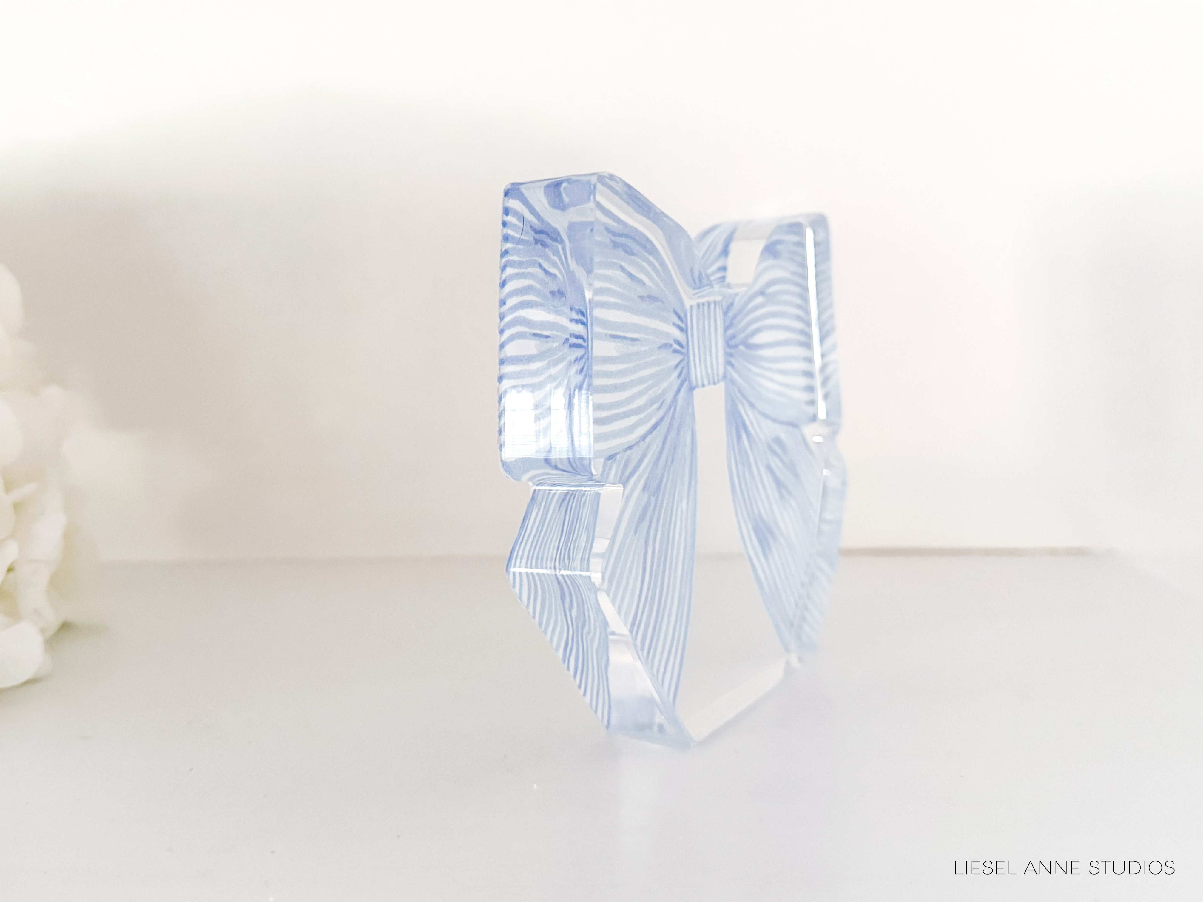 A clear glass object with a blue and white striped pattern, resembling a bow or ribbon, is displayed on a white surface.