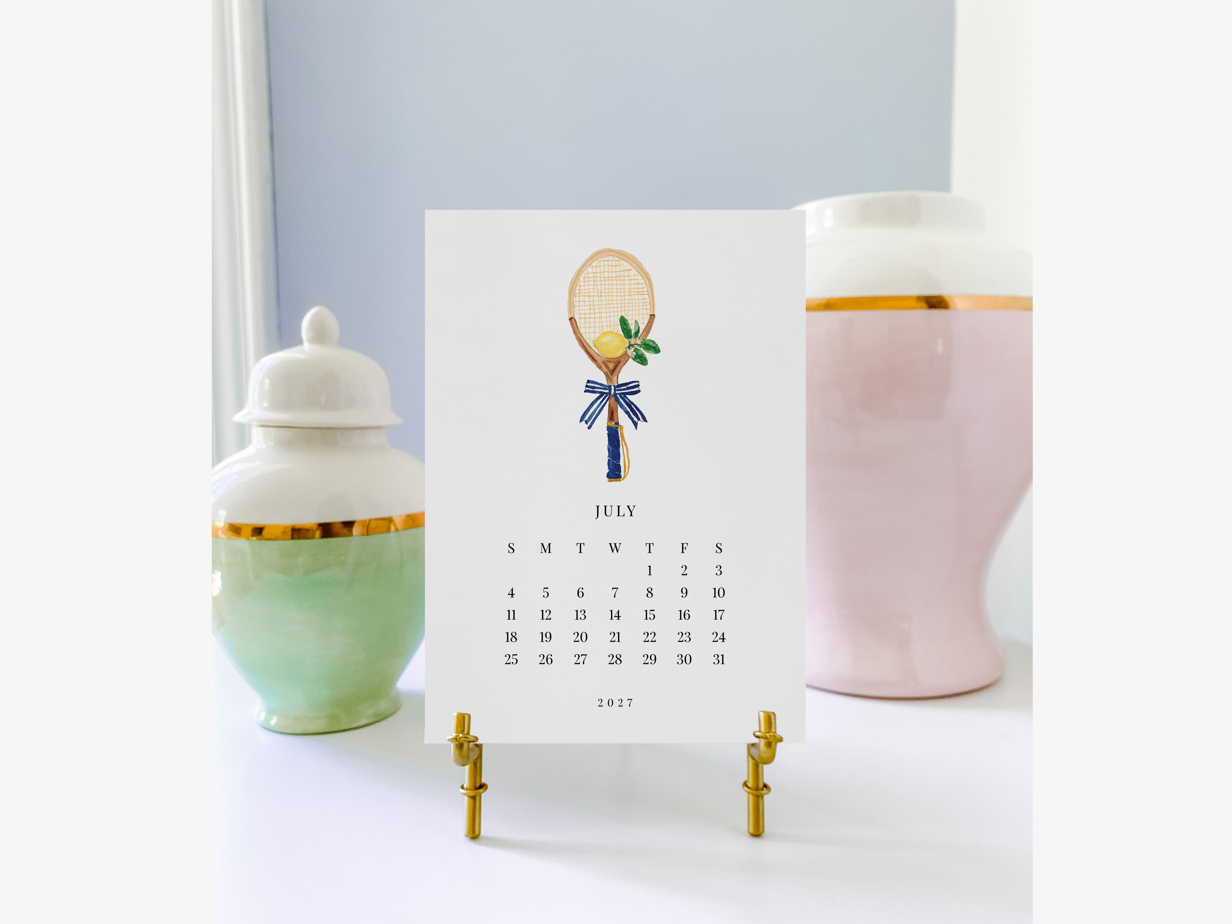 A white calendar with a tennis racket illustration on it, placed on a stand in front of two decorative vases.