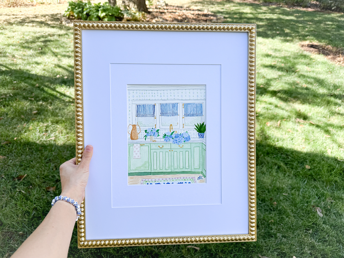 A hand is holding a framed picture of a kitchen scene with a green cabinet, blue countertop, and various kitchen items such as a vase and a potted plant.