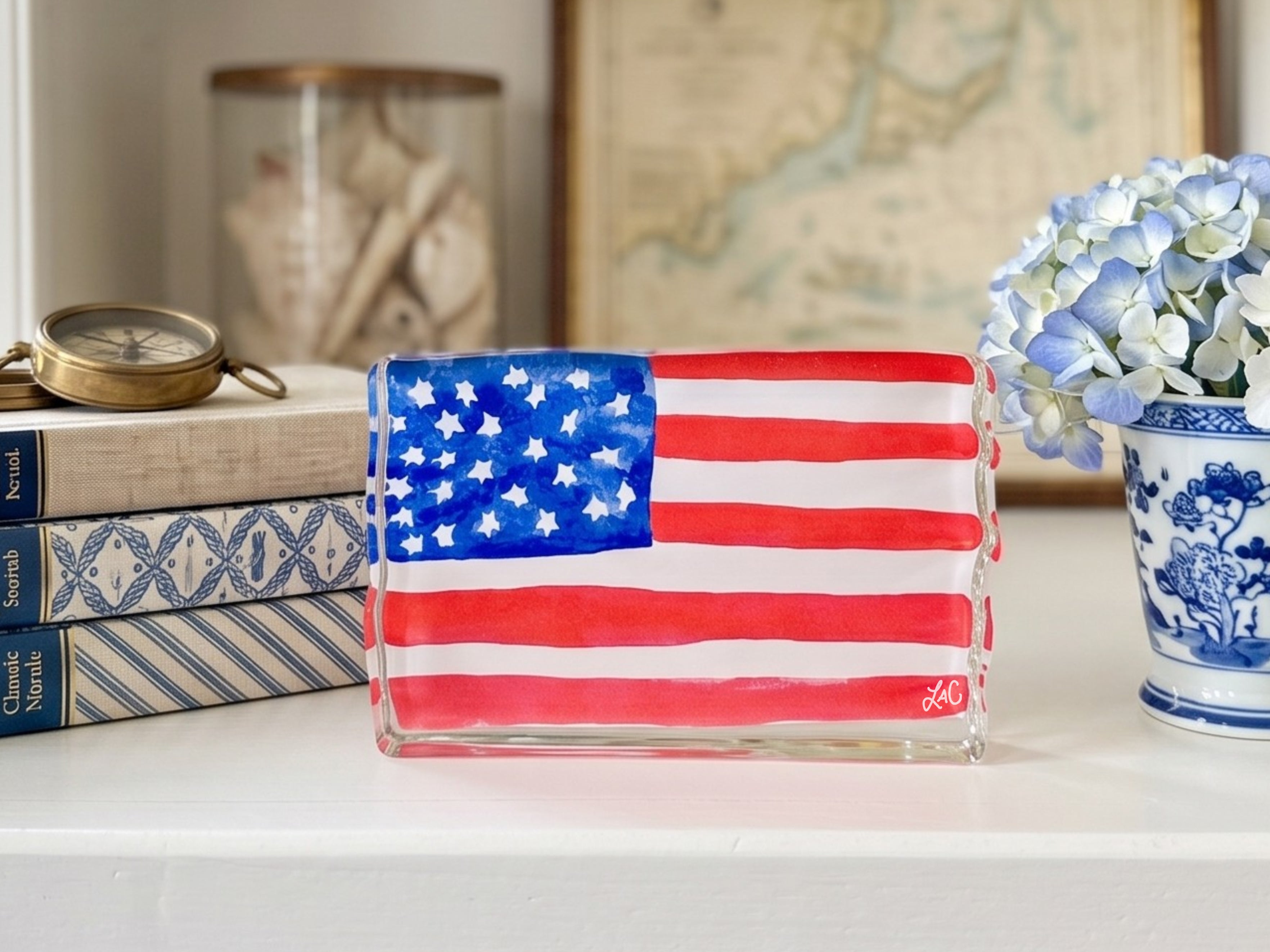 a white shelf with various objects, including a clear glass sculpture of the American flag, a blue and white vase with blue and white flowers, and a stack of books.