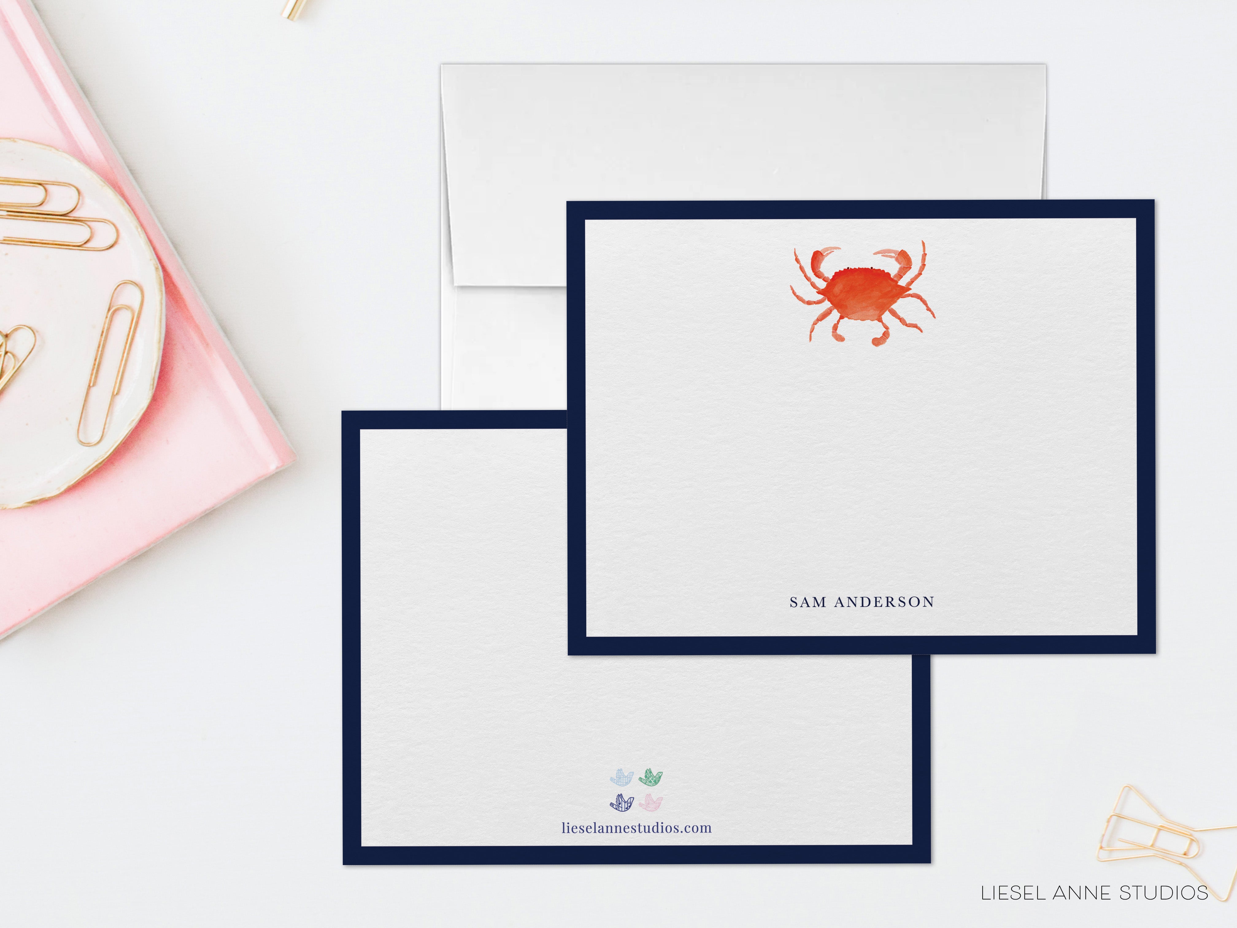 two blank stationery cards with a watercolor illustration of a crab on them, placed on a white surface alongside a pink object and a white envelope.