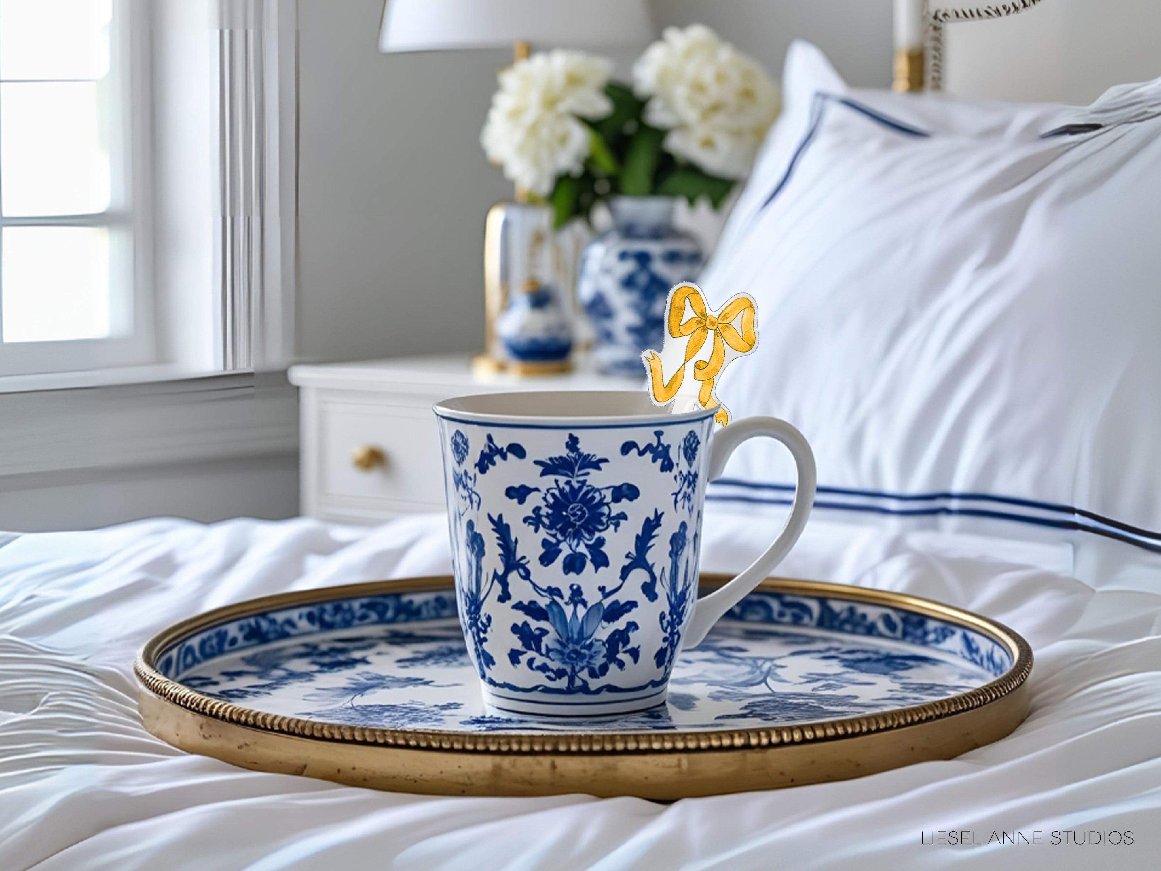 A blue and white patterned coffee mug with a gold rim sits on a white tray, placed on a bed with white sheets and a white pillow.