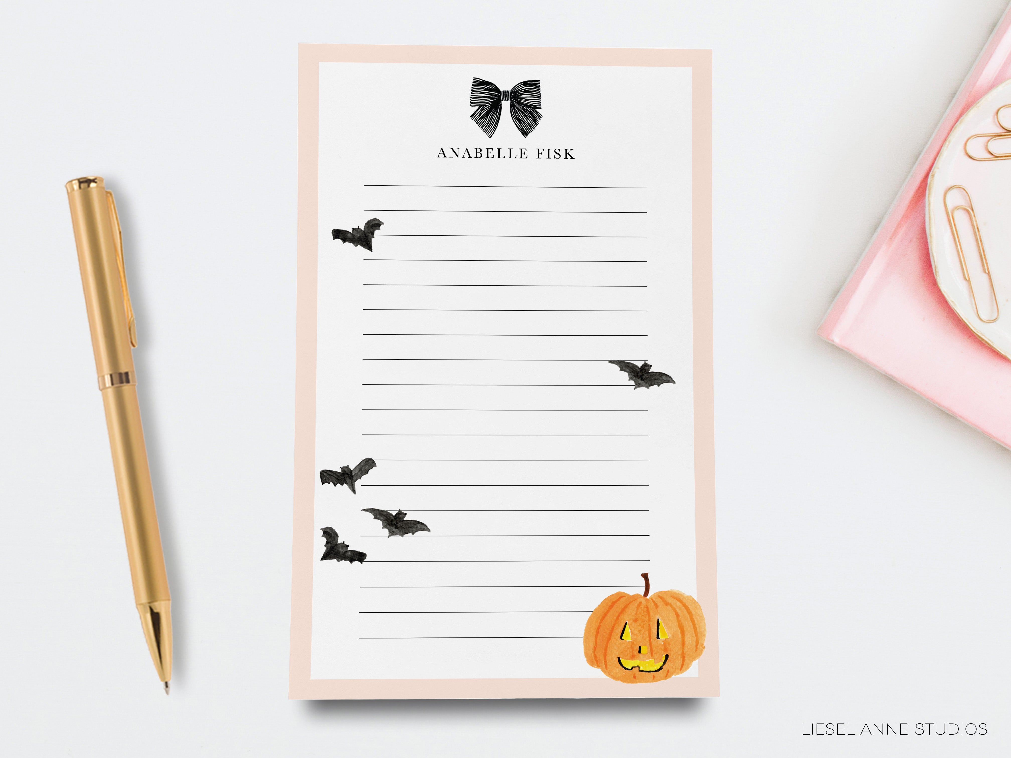a pink sticky note pad with a black bat illustration, a gold pen, and a small pumpkin decoration.