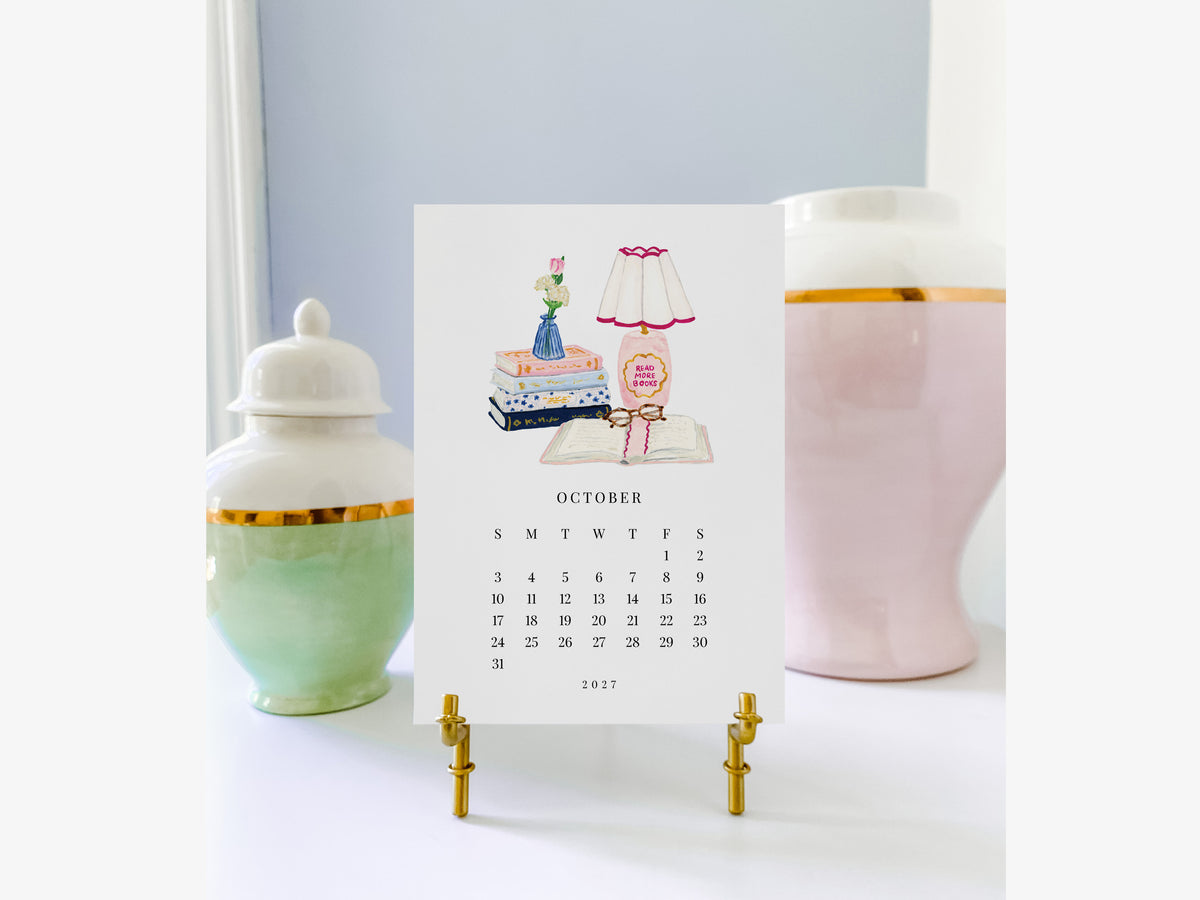 A calendar for the month of October 2017, featuring illustrations of a lamp, books, and a vase, placed on a white surface with two decorative vases in the background.