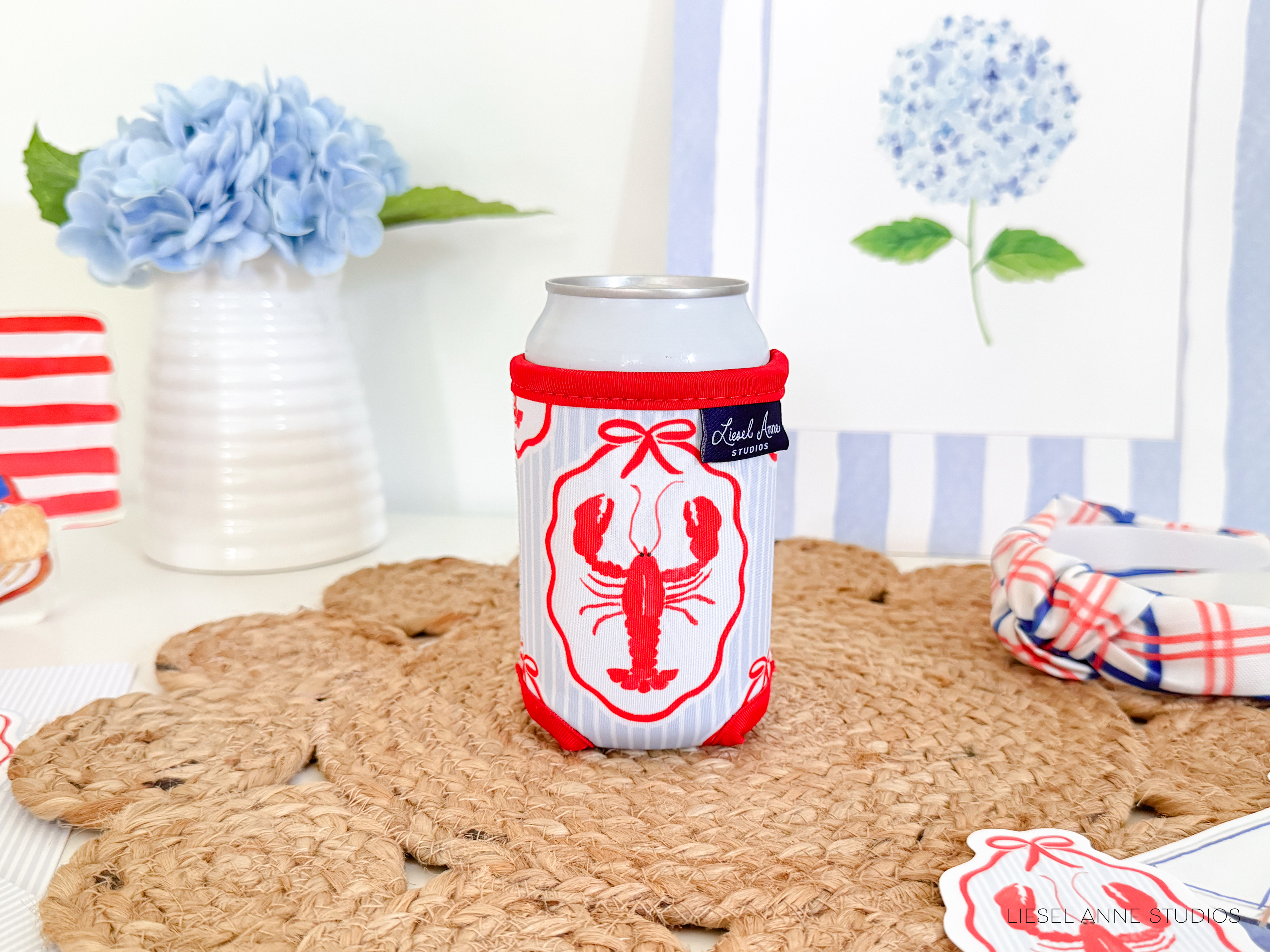A can of soda with a lobster design sits on a woven placemat, surrounded by a vase of blue hydrangeas, a striped coaster, and a framed picture of a blue hydrangea.