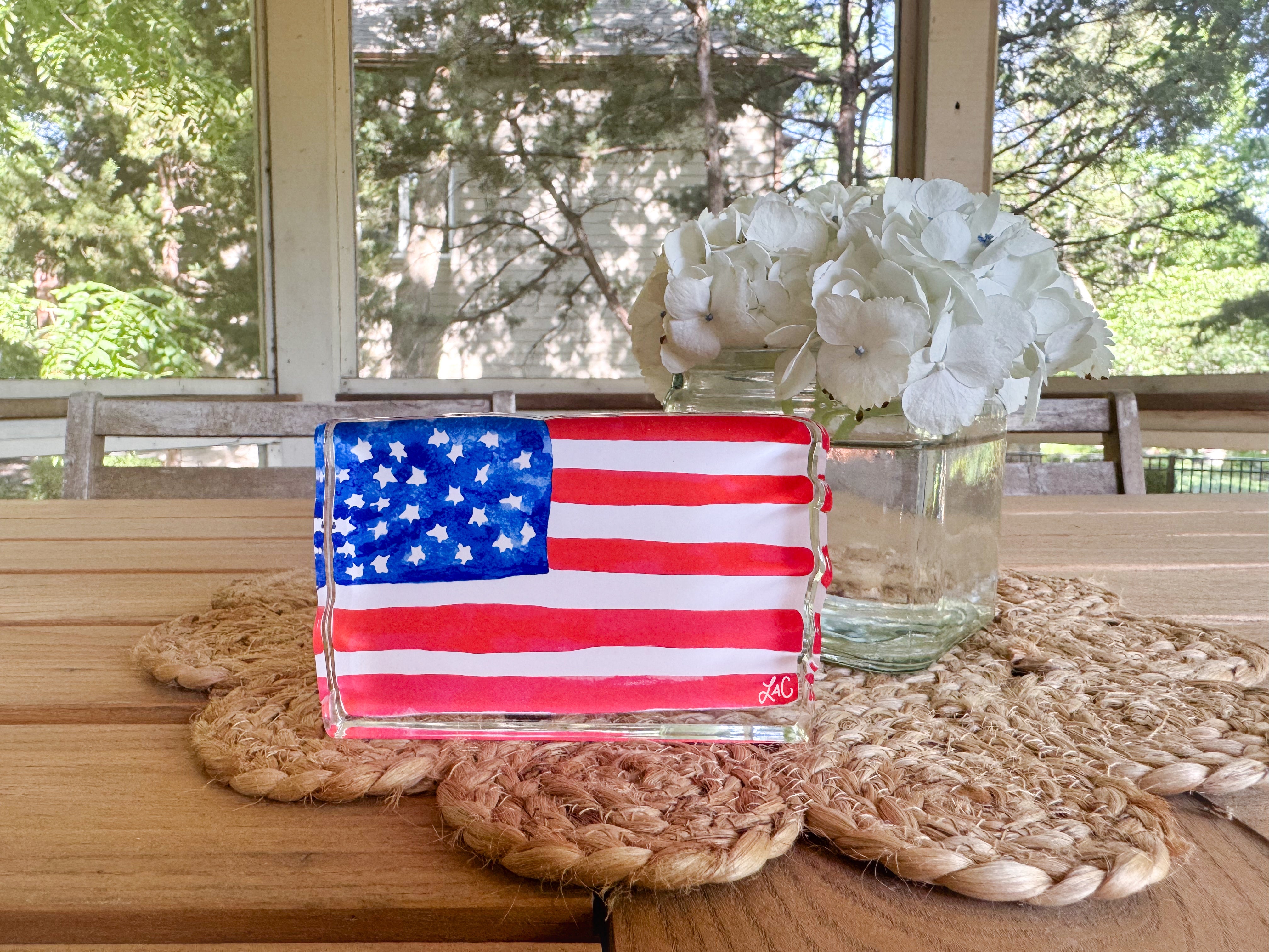 a wooden table with a clear glass vase containing white hydrangeas, a small American flag painted on a glass block, and a woven placemat.