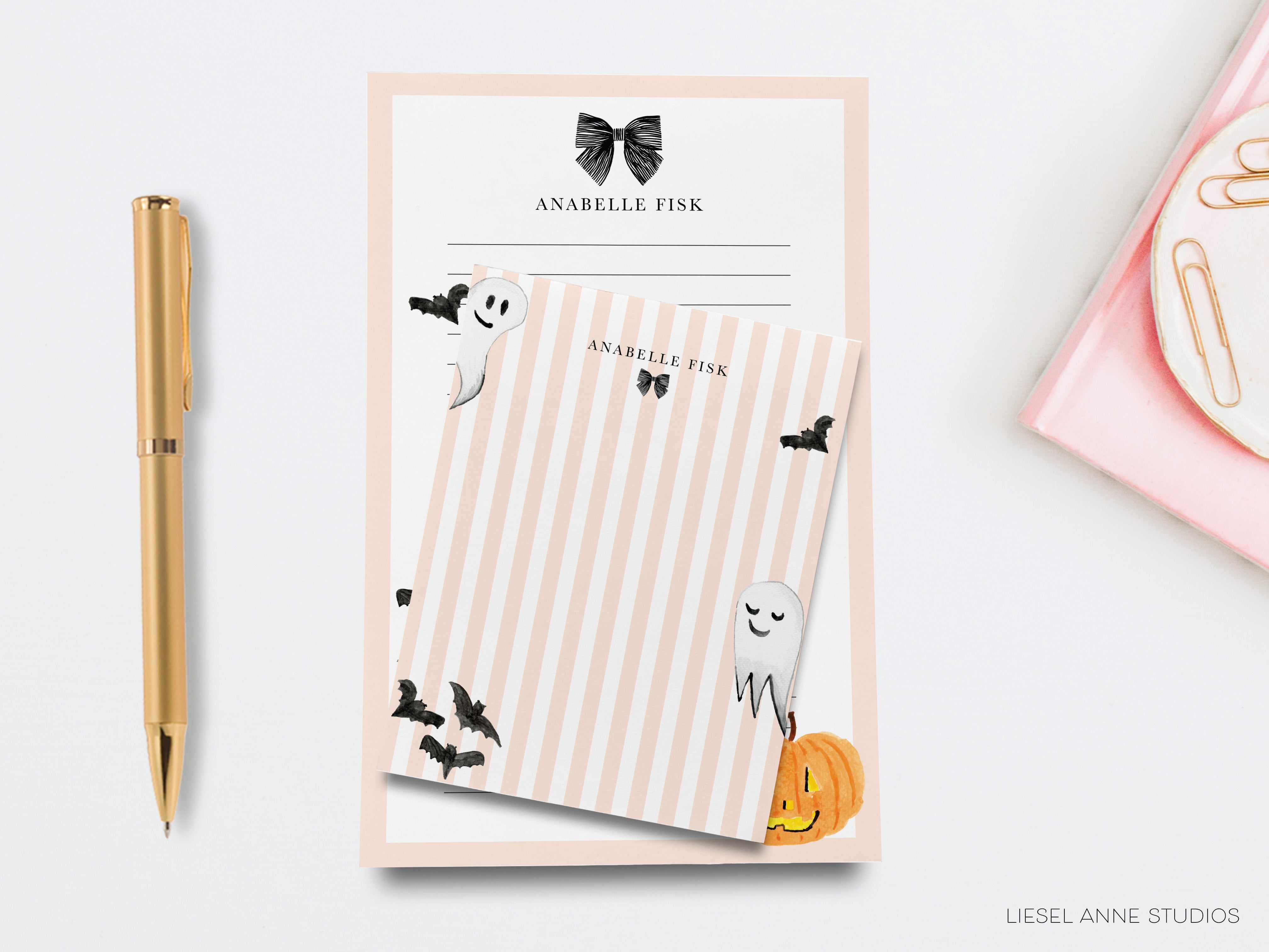 a pink and white striped notepad with a Halloween-themed design, including a ghost, bats, and a pumpkin. There is also a gold pen and a pink object on the right side of the image.