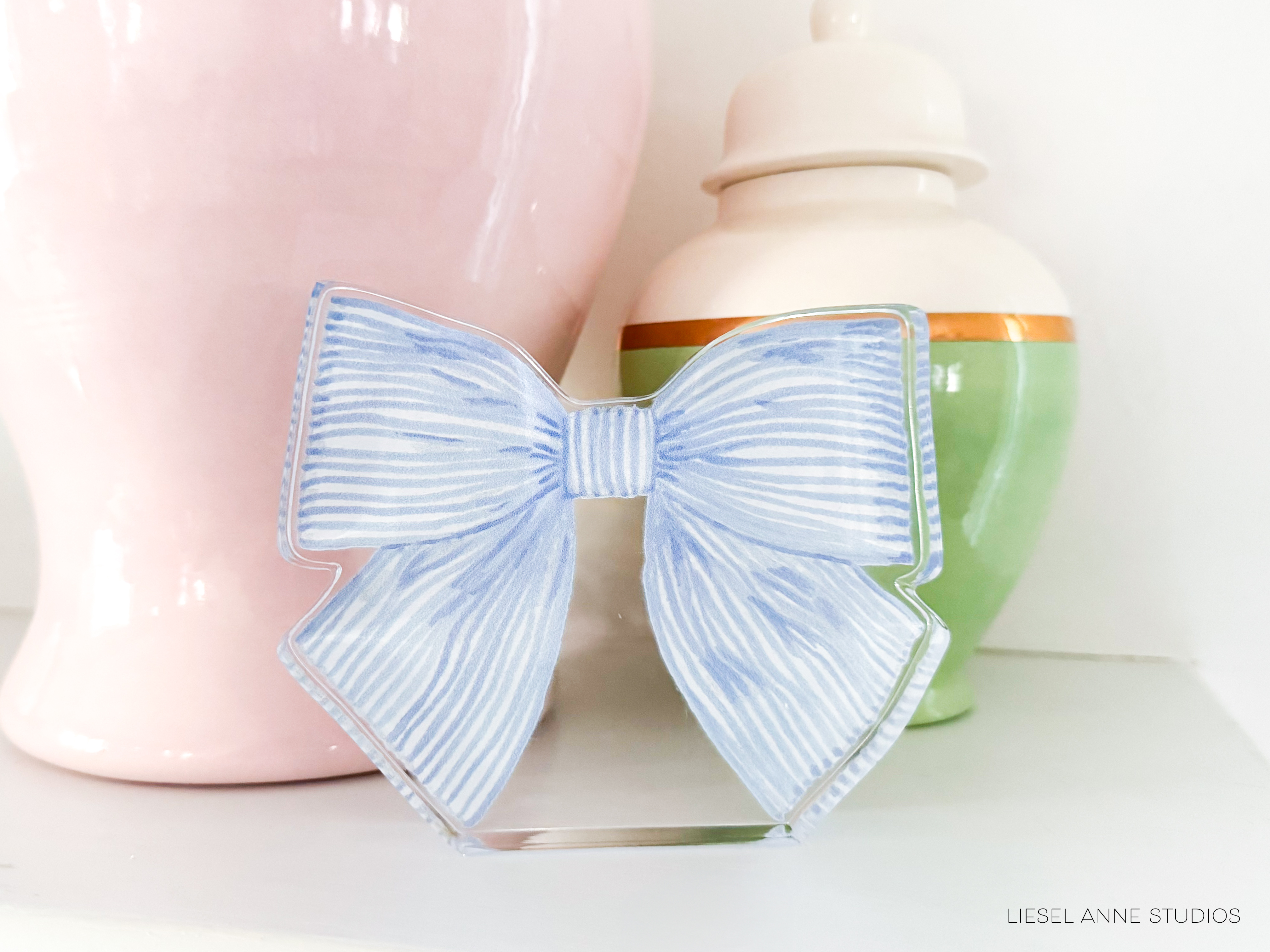 A blue and white striped bow is displayed on a shelf, accompanied by a pink vase and a green vase.