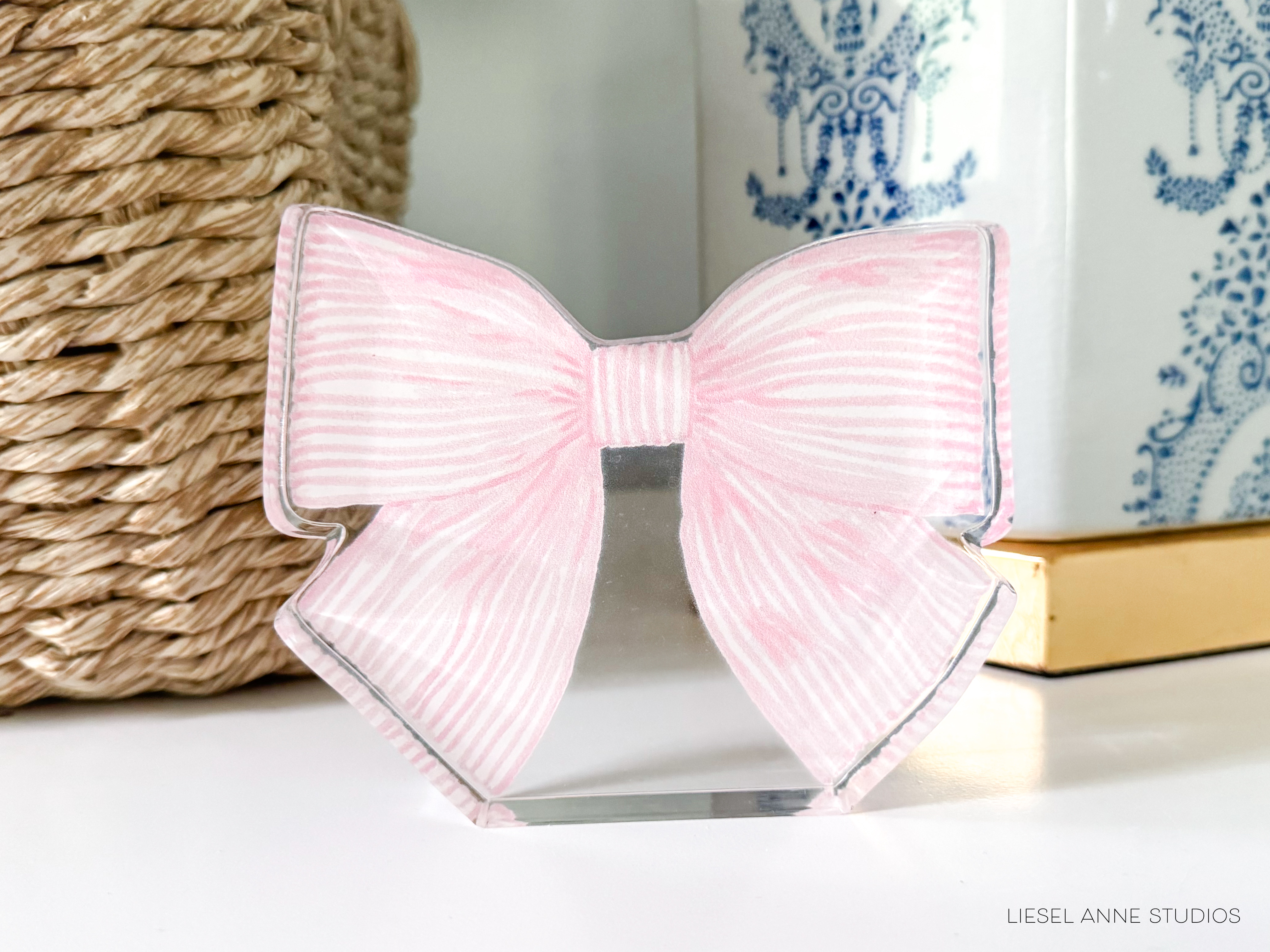 A pink and white striped bow-shaped object is displayed on a white surface, with a woven basket and a blue and white patterned vase in the background.