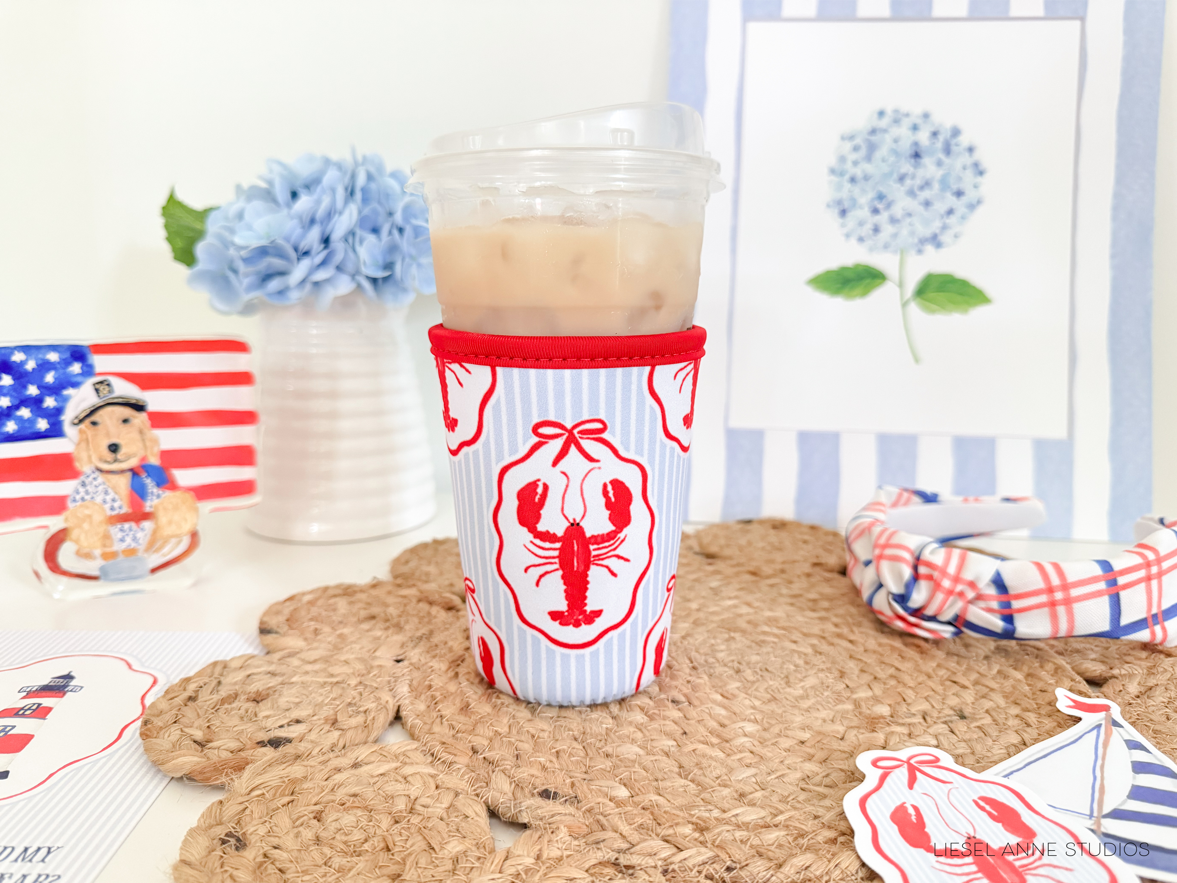 a cup with a red and white striped design, placed on a woven placemat, and a cup with a red and white striped design, placed on a woven placemat. The background includes a framed picture of a blue flower, a small American flag, and a stuffed animal.