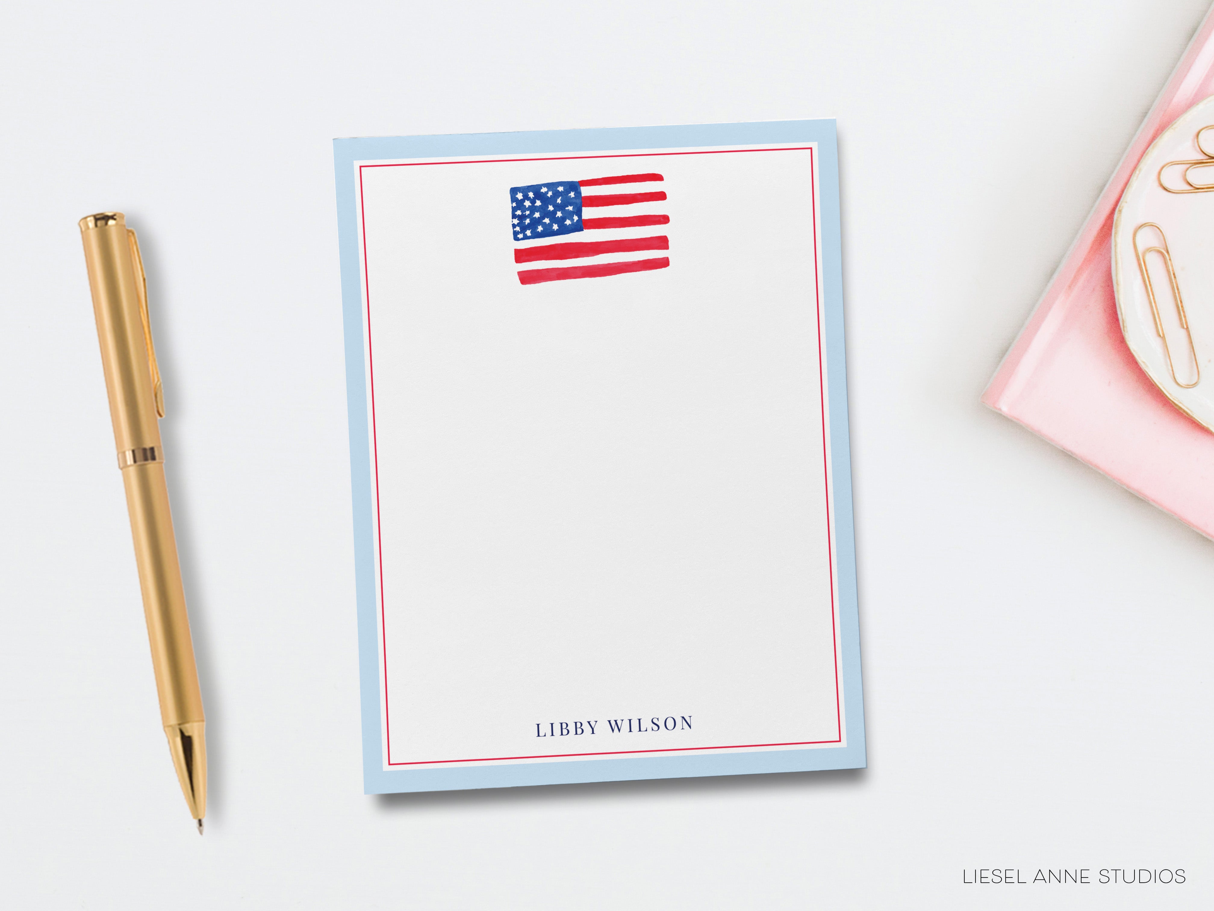a blue notepad with an American flag design, a gold pen, and a pink object, all placed on a white surface.