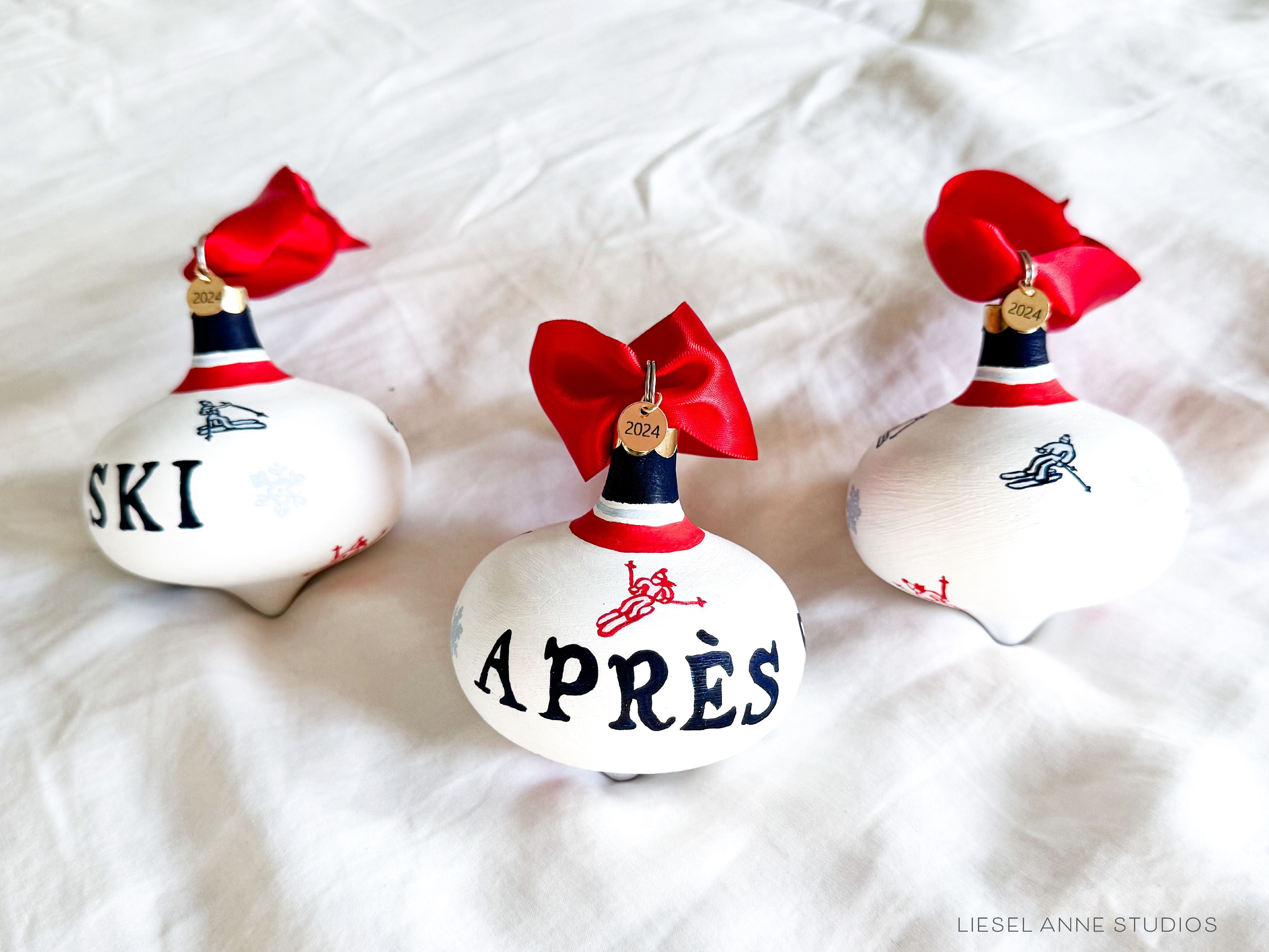 Après Ski Hand-Painted Ceramic Bauble-Ceramic Hand-Painted Keepsake Ornament, Measures 3" x 3", Features the red, blue & white preppy skier design with a white base, a gold scalloped cap, and a beautiful thick satin red ribbon ready for hanging on your tree, Includes a gold charm with the year 2024 on the front and the Liesel Anne Studios birds on the back, Coated with a protective matte finish, Each bauble is slightly different with potential small imperfections due to the hand-painted nature, making e