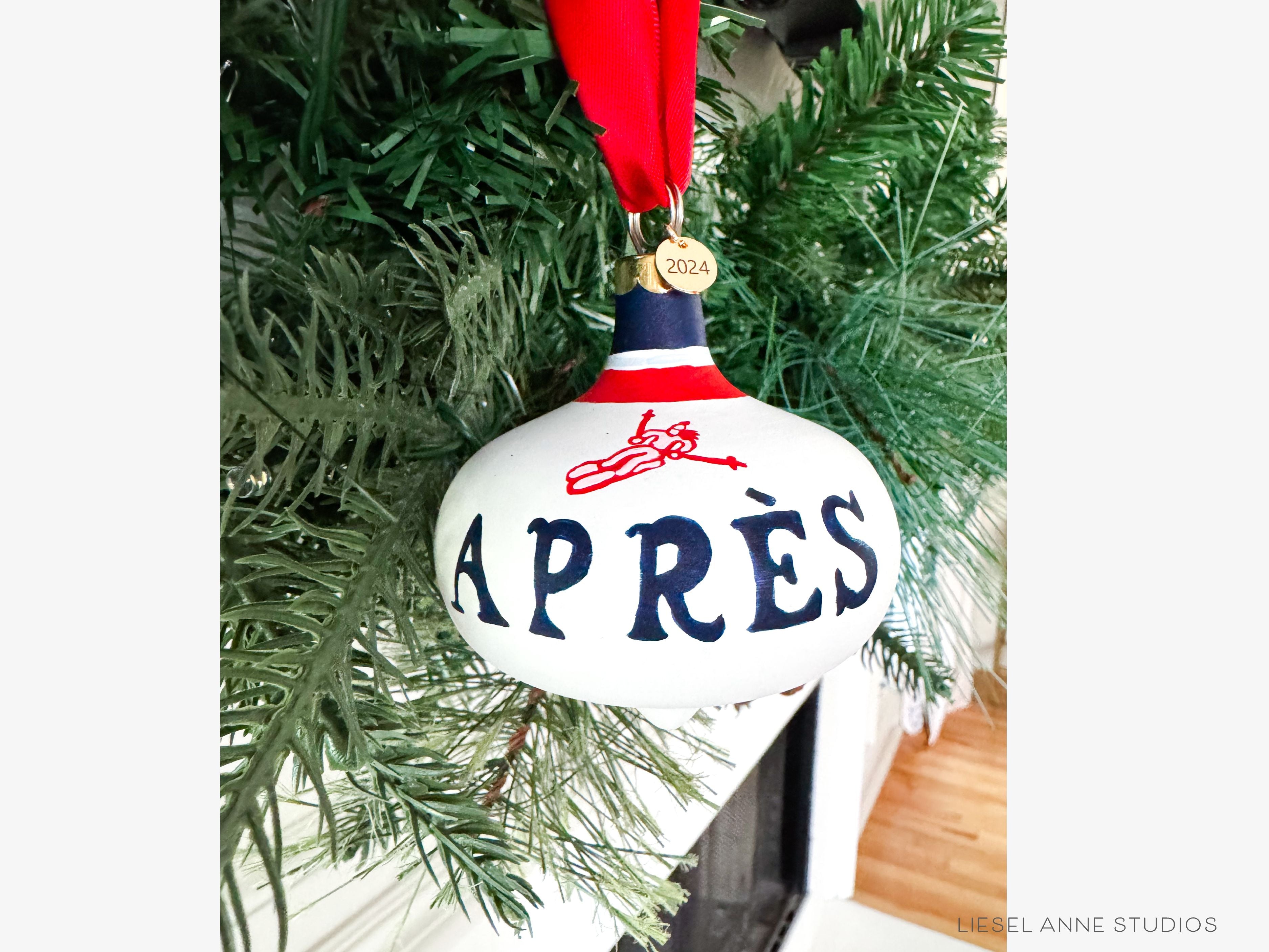 Après Ski Hand-Painted Ceramic Bauble-Ceramic Hand-Painted Keepsake Ornament, Measures 3" x 3", Features the red, blue & white preppy skier design with a white base, a gold scalloped cap, and a beautiful thick satin red ribbon ready for hanging on your tree, Includes a gold charm with the year 2024 on the front and the Liesel Anne Studios birds on the back, Coated with a protective matte finish, Each bauble is slightly different with potential small imperfections due to the hand-painted nature, making e