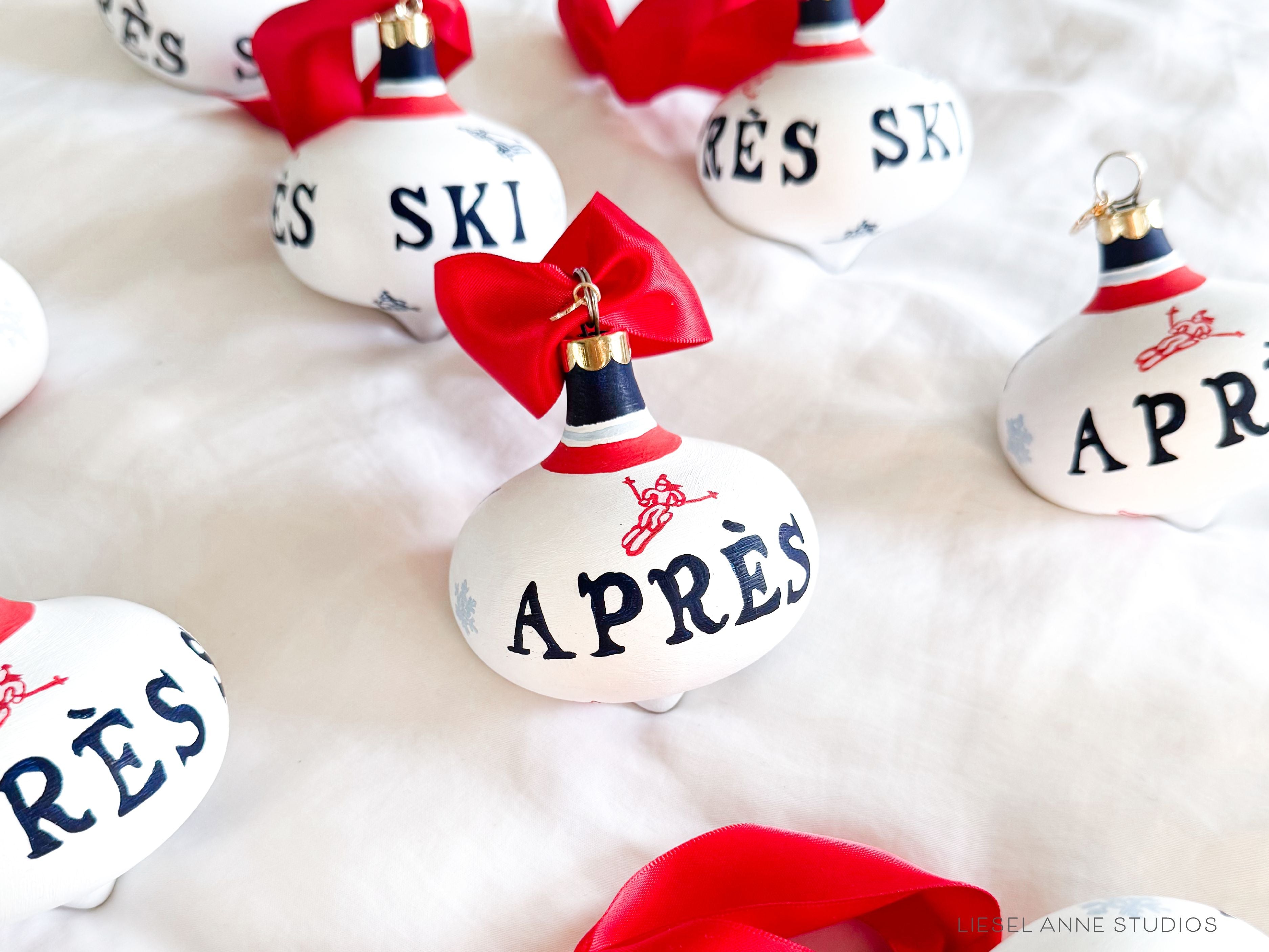 Après Ski Hand-Painted Ceramic Bauble-Ceramic Hand-Painted Keepsake Ornament, Measures 3" x 3", Features the red, blue & white preppy skier design with a white base, a gold scalloped cap, and a beautiful thick satin red ribbon ready for hanging on your tree, Includes a gold charm with the year 2024 on the front and the Liesel Anne Studios birds on the back, Coated with a protective matte finish, Each bauble is slightly different with potential small imperfections due to the hand-painted nature, making e