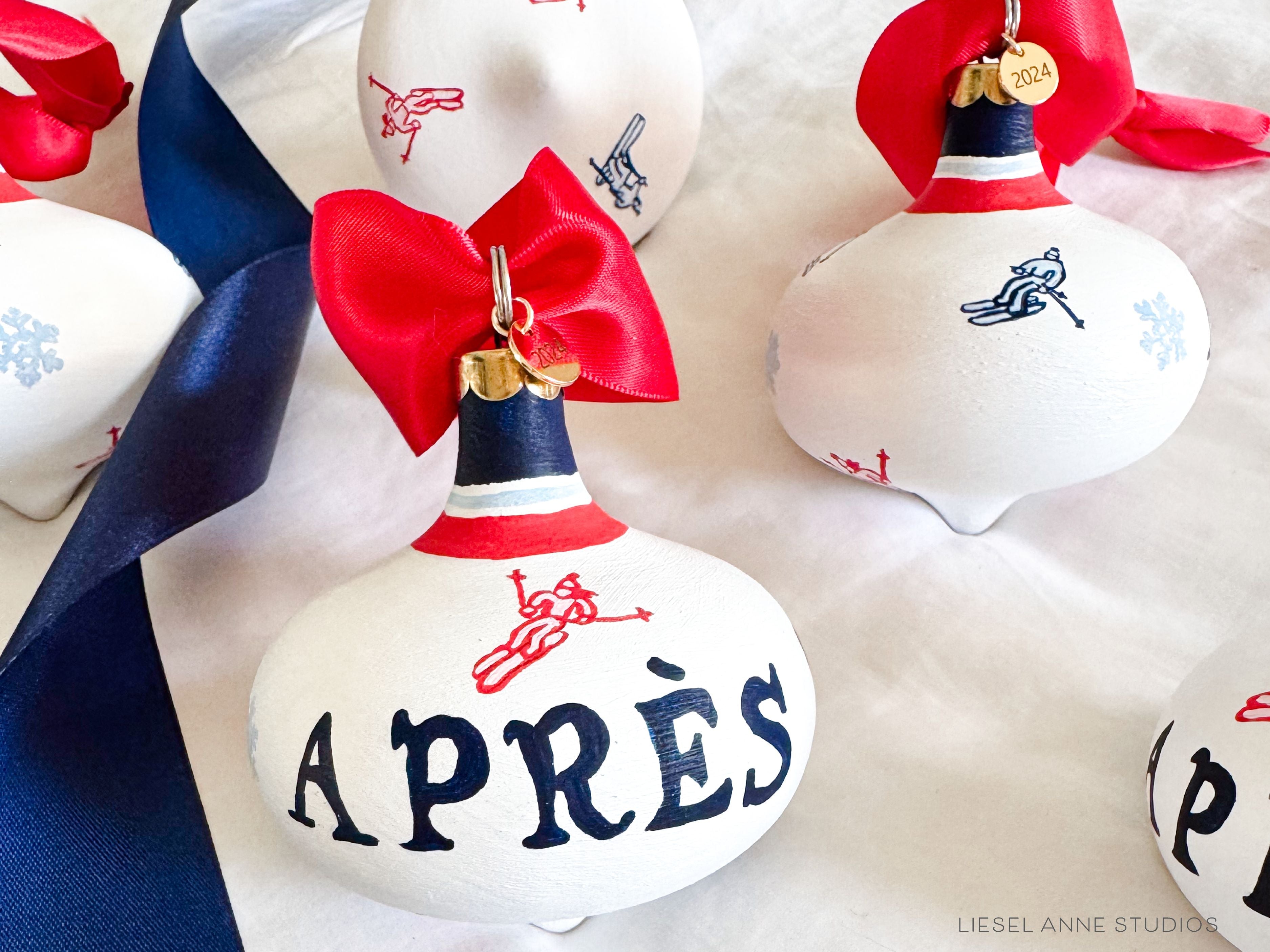Après Ski Hand-Painted Ceramic Bauble-Ceramic Hand-Painted Keepsake Ornament, Measures 3" x 3", Features the red, blue & white preppy skier design with a white base, a gold scalloped cap, and a beautiful thick satin red ribbon ready for hanging on your tree, Includes a gold charm with the year 2024 on the front and the Liesel Anne Studios birds on the back, Coated with a protective matte finish, Each bauble is slightly different with potential small imperfections due to the hand-painted nature, making e
