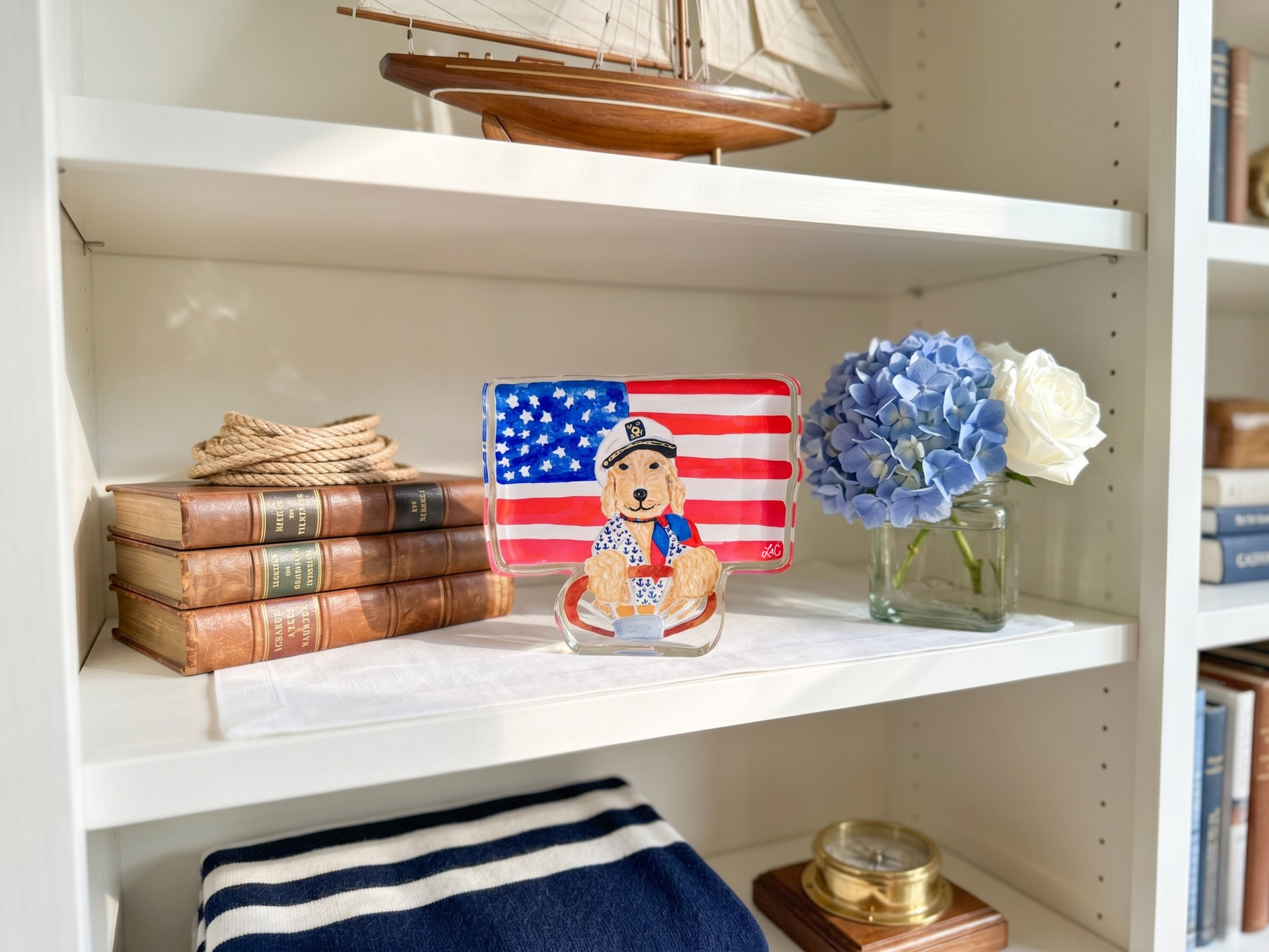 a bookshelf with various items, including a small American flag, a model ship, a blue hydrangea flower, and a stack of books.