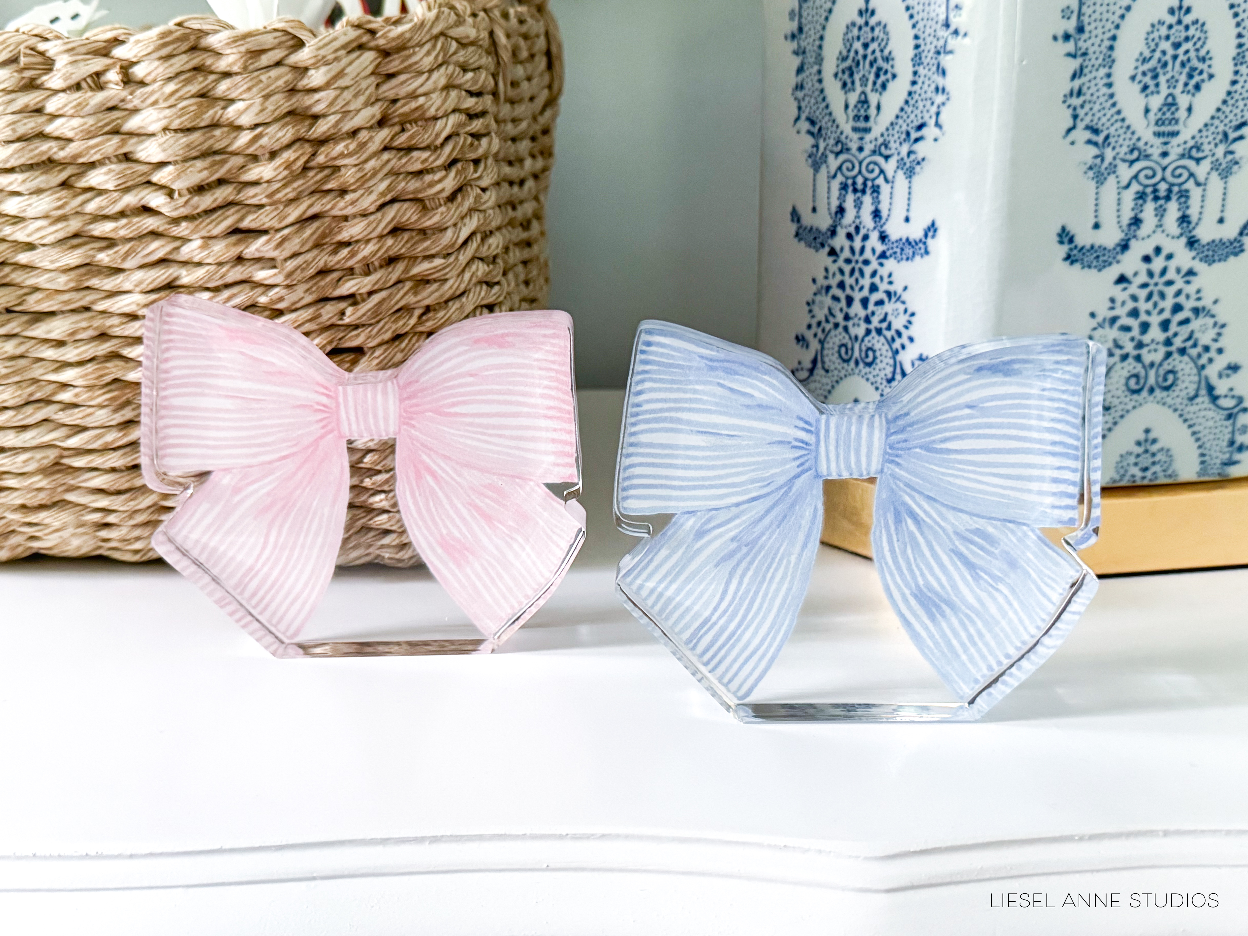 two decorative bow ties, one pink and one blue, placed on a white surface. There is also a woven basket and a blue and white patterned vase in the background.