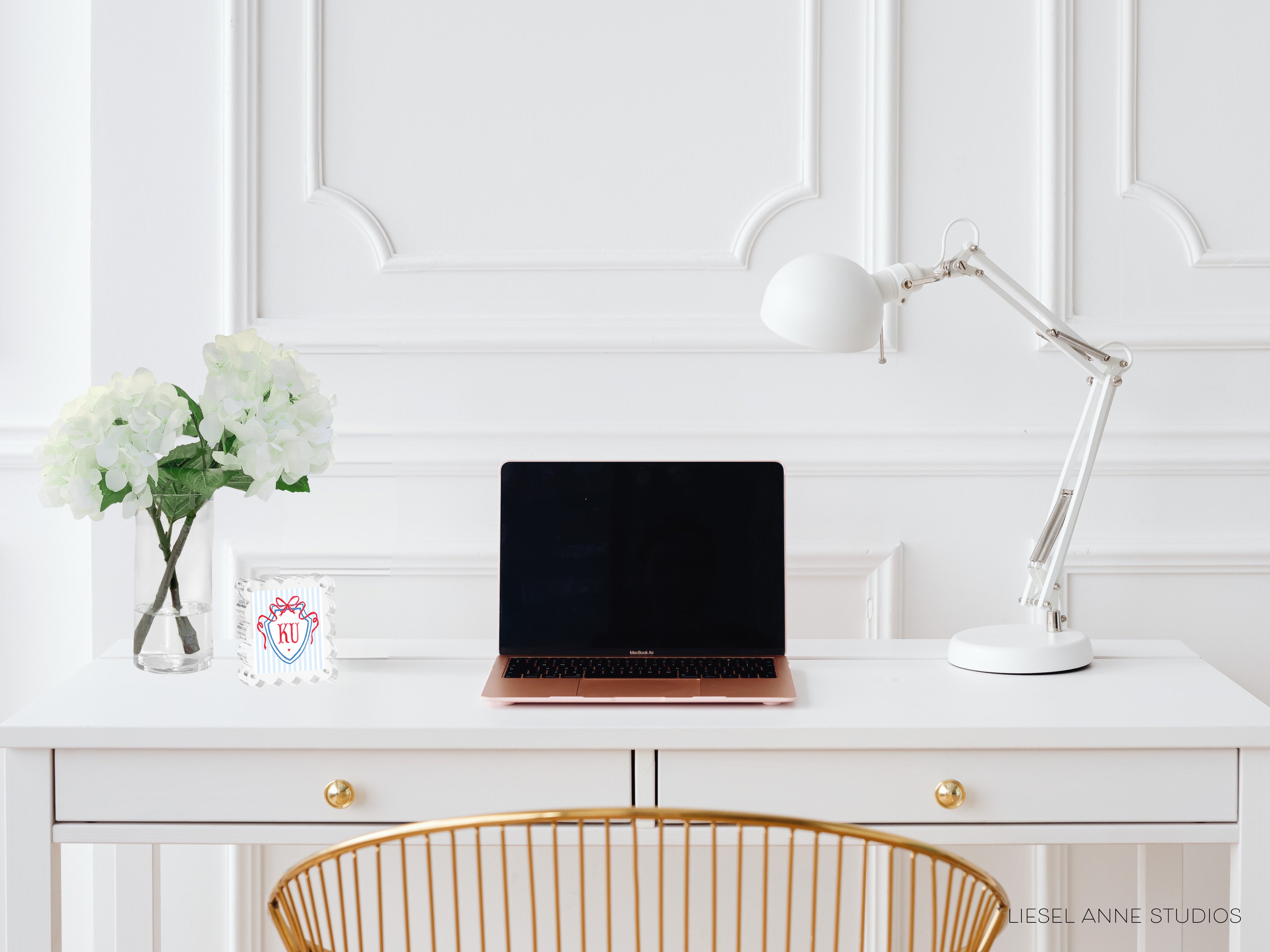 A white desk with a laptop, a vase of white flowers, and a white lamp, all arranged neatly and creating a clean, minimalist workspace.