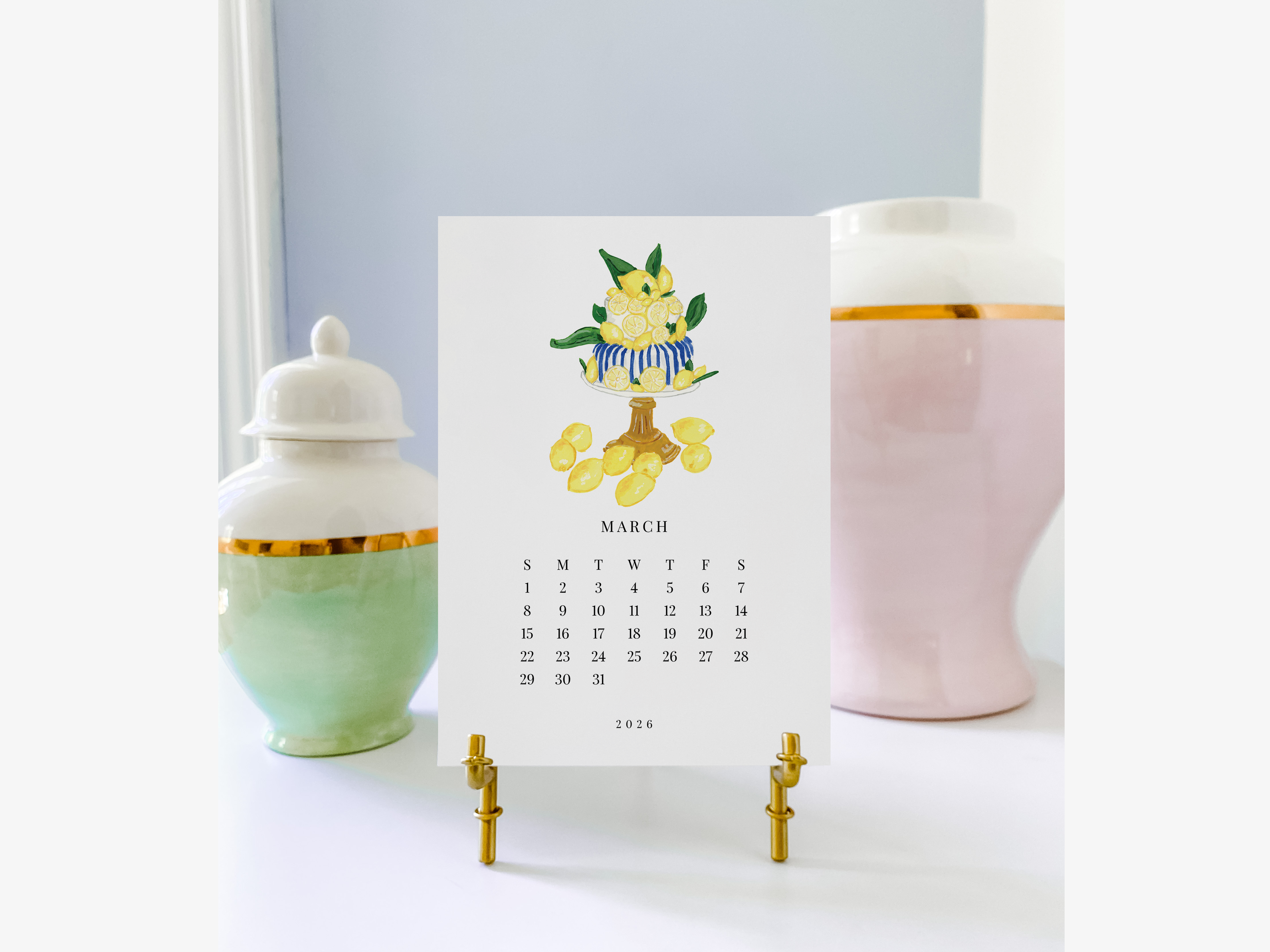 A calendar for the month of March 2016, featuring a lemon illustration, is displayed on a stand between two ceramic vases.